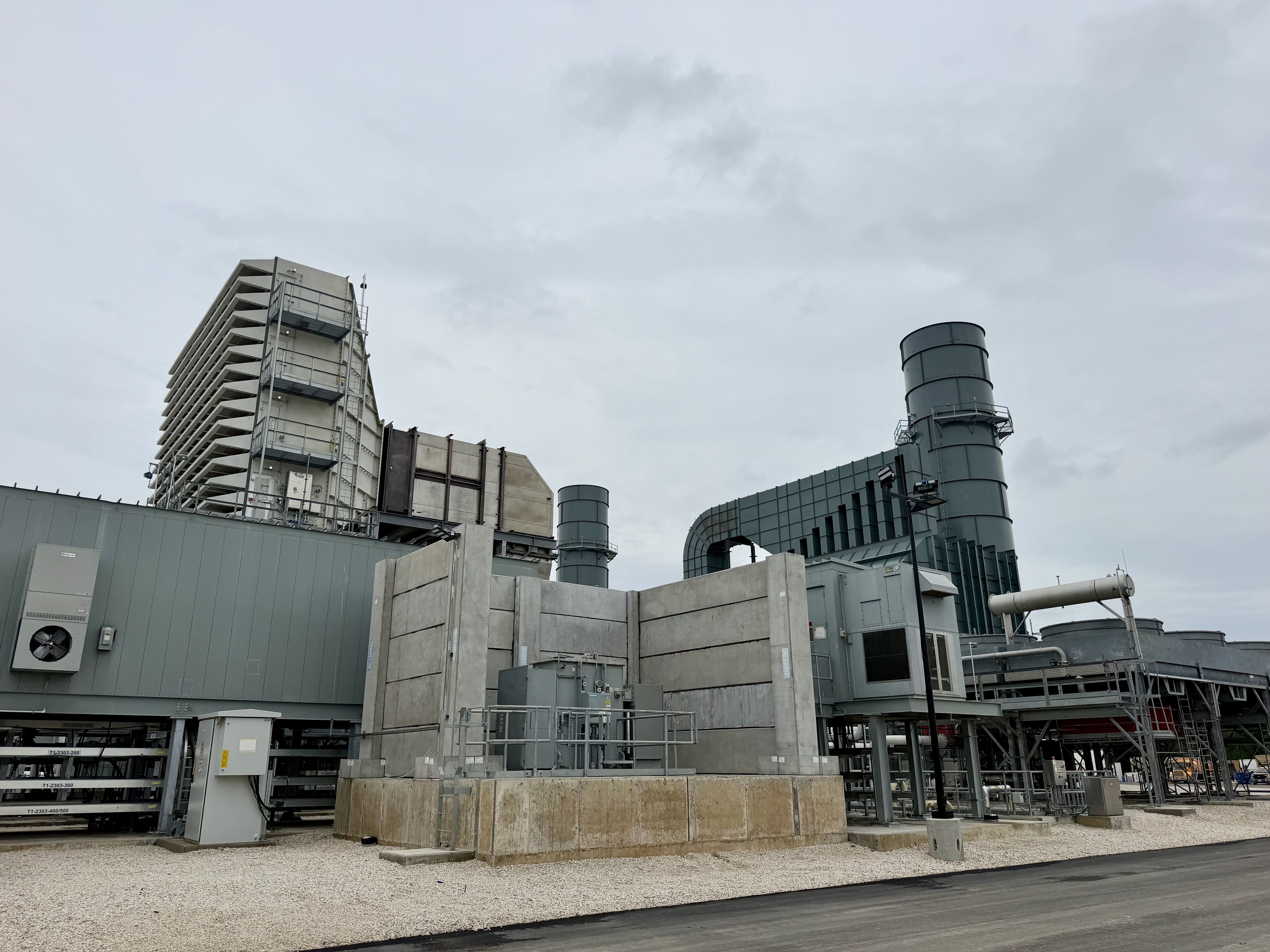 Industrial facility with gray modular buildings, a tall stairwell structure, large cylindrical vents, and interconnected piping. Gravel ground beneath a cloudy blue sky.