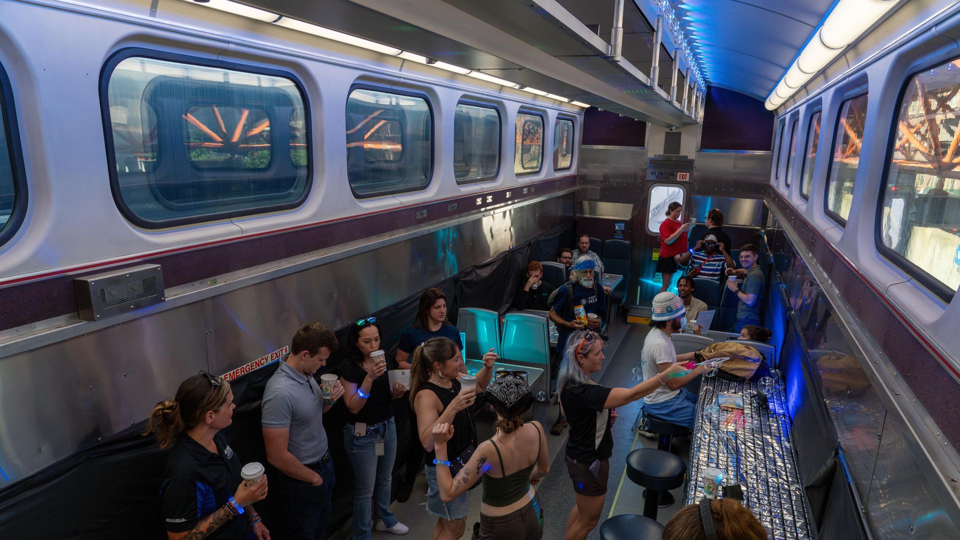People socializing between two trains with large windows, some standing with drinks, others seated. Blue LED lights illuminate the ceiling and a bar with stools lines one wall.