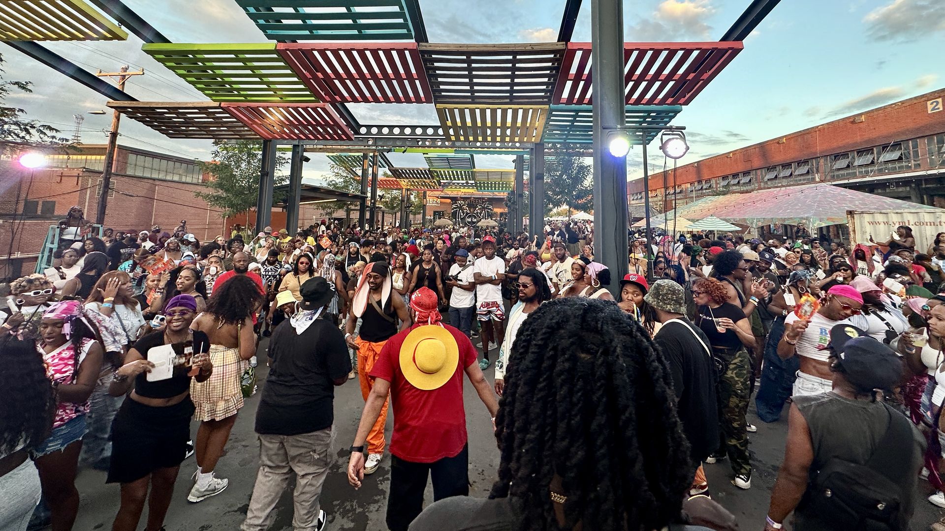 Large crowd of people dressed casually in bright colors gathered outdoors under artistic colorful slatted canopies on a sunny day with blue sky and scattered clouds.
