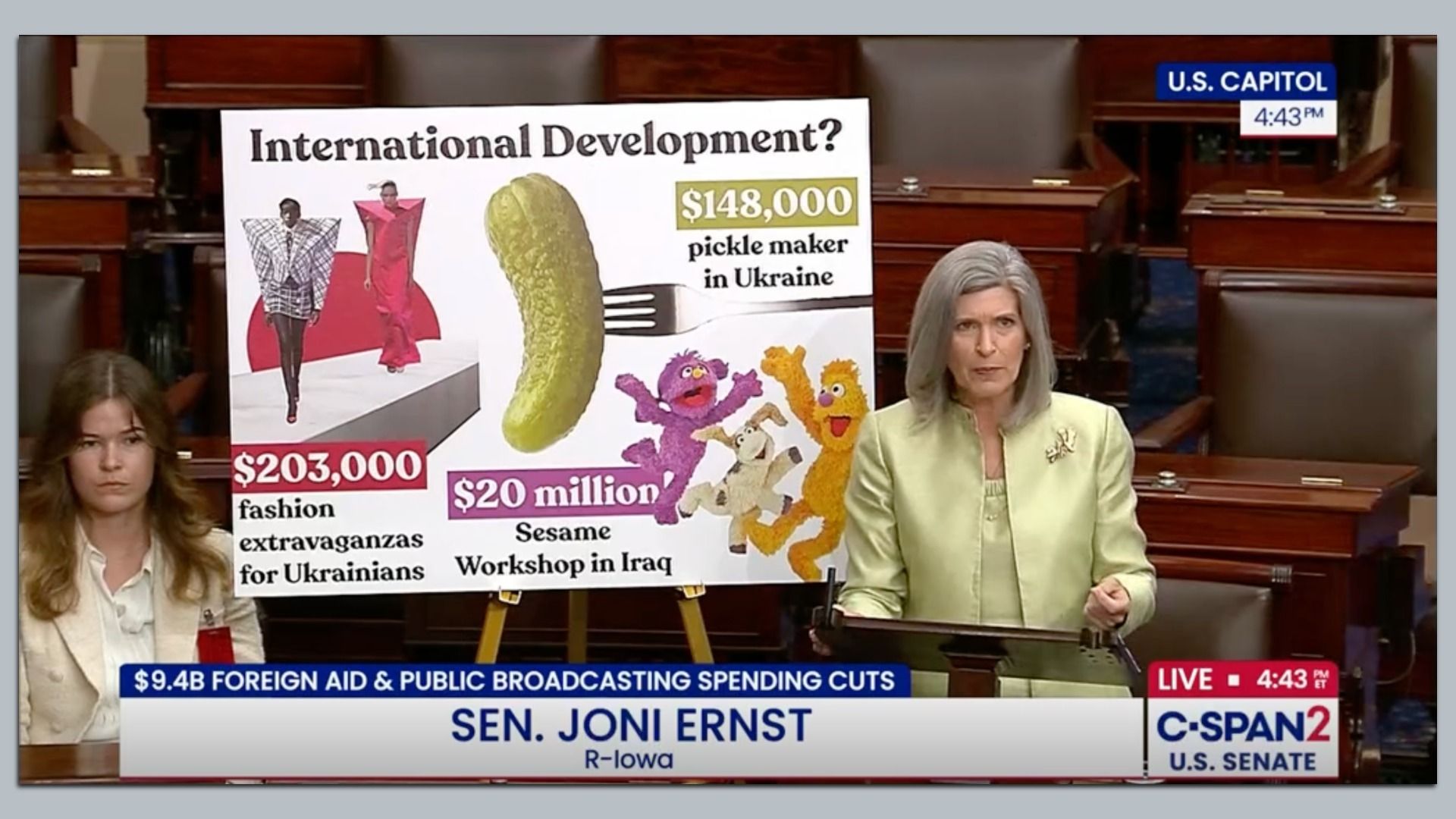 Sen. Joni Ernst at U.S. Senate podium with a poster on international development showing $203,000 for Ukrainian fashion, $148,000 for a pickle maker in Ukraine, and $20 million for Sesame Workshop in Iraq.