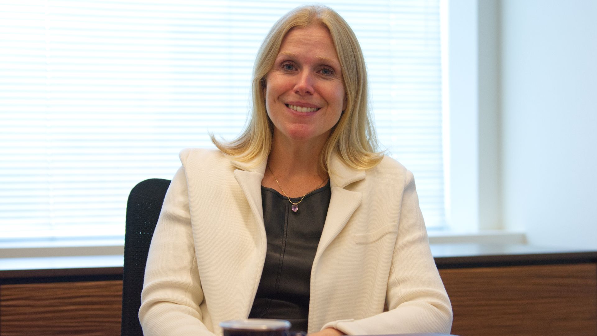 Lauren Knausenberger, a woman, sits a desk in a well-lit office. She is wearing a black shirt and white blazer.