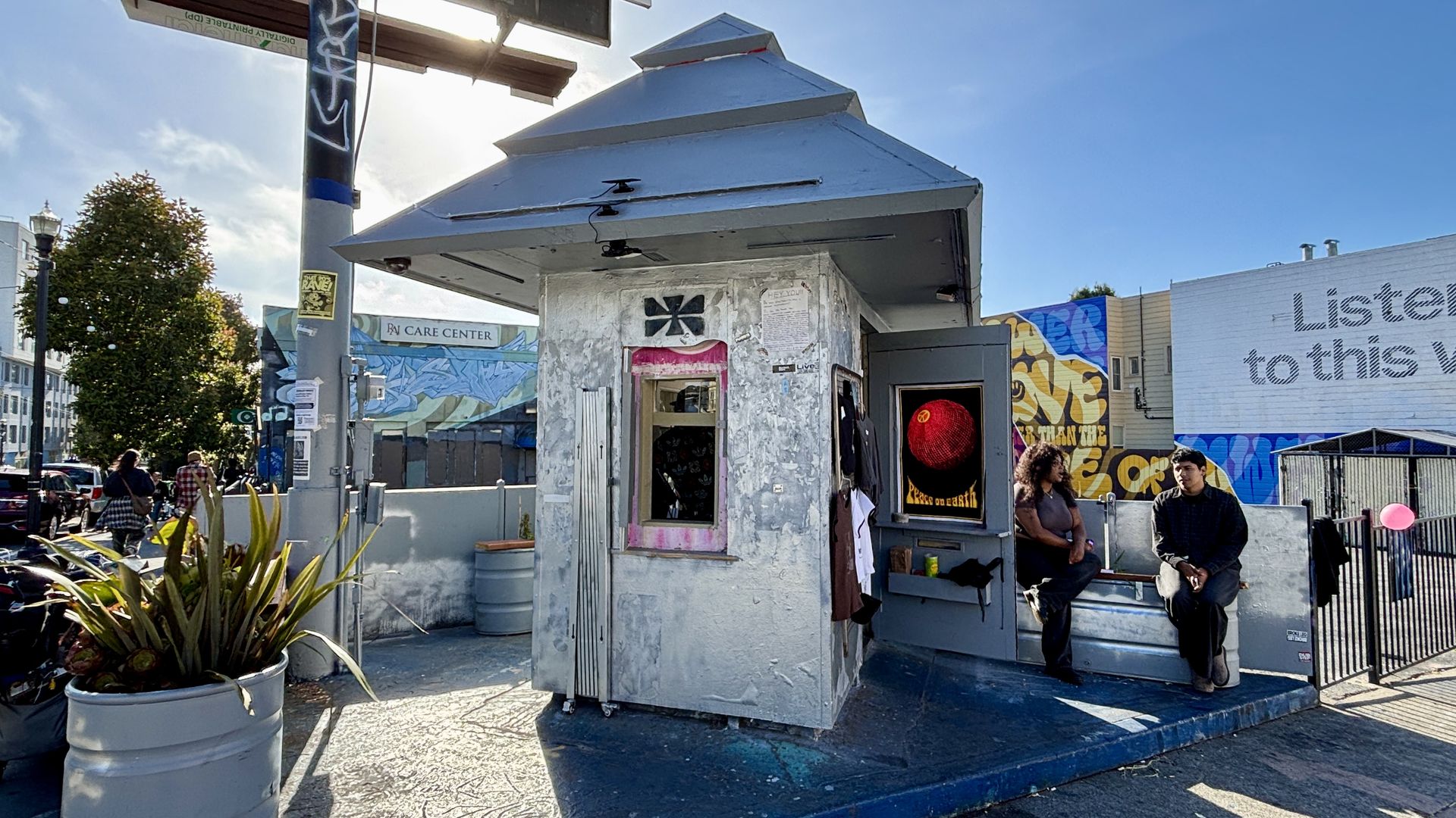 Weathered silver kiosk with a slanted roof on a sunny plaza; posters and a window, graffiti walls behind; two casually dressed men stand nearby; a tree and planters frame the left.