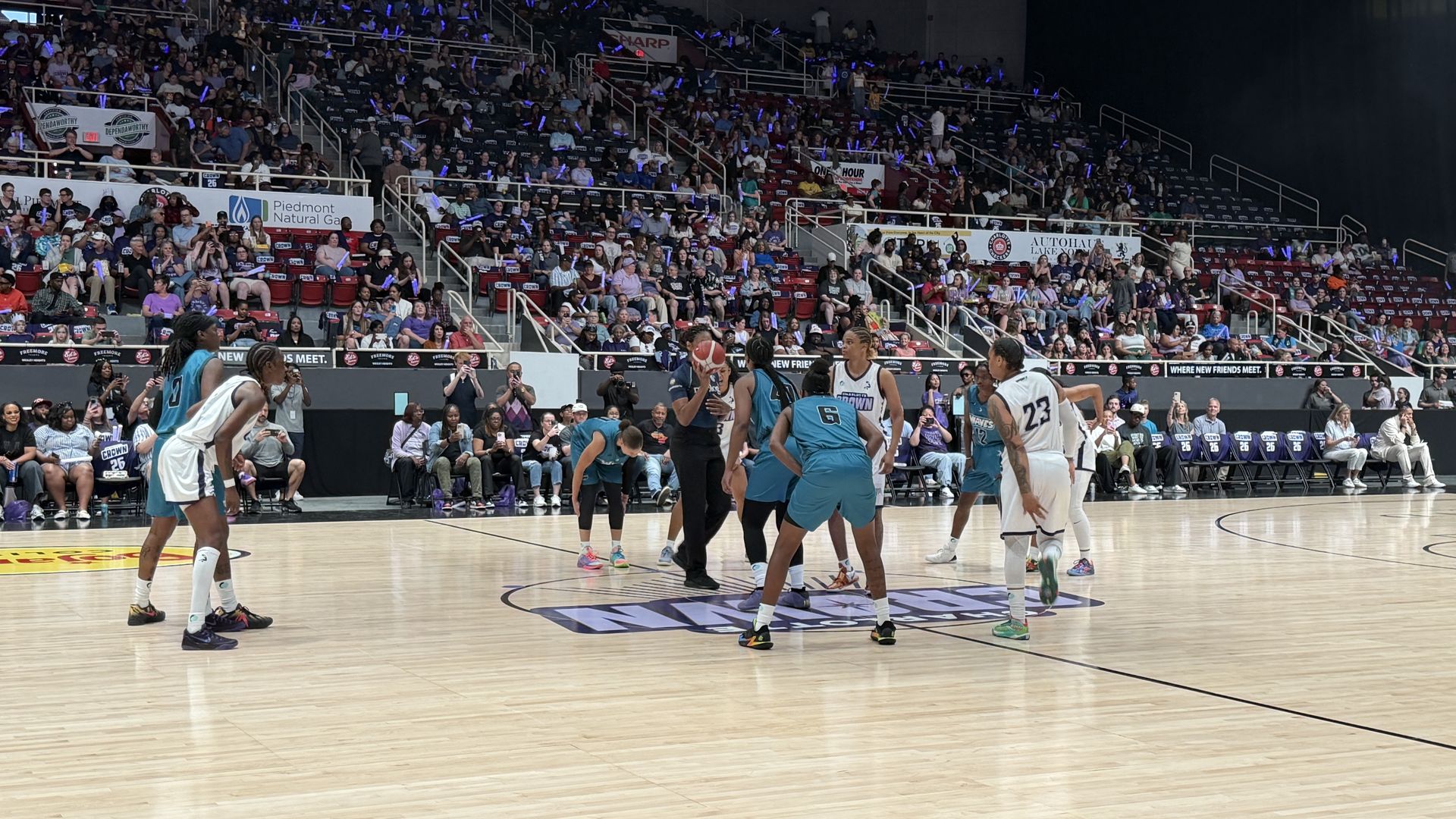 Basketball game in a crowded arena: players in teal and white battle for the ball on a polished court, as a large audience with blue glow sticks, banners, and signs fills the stands.