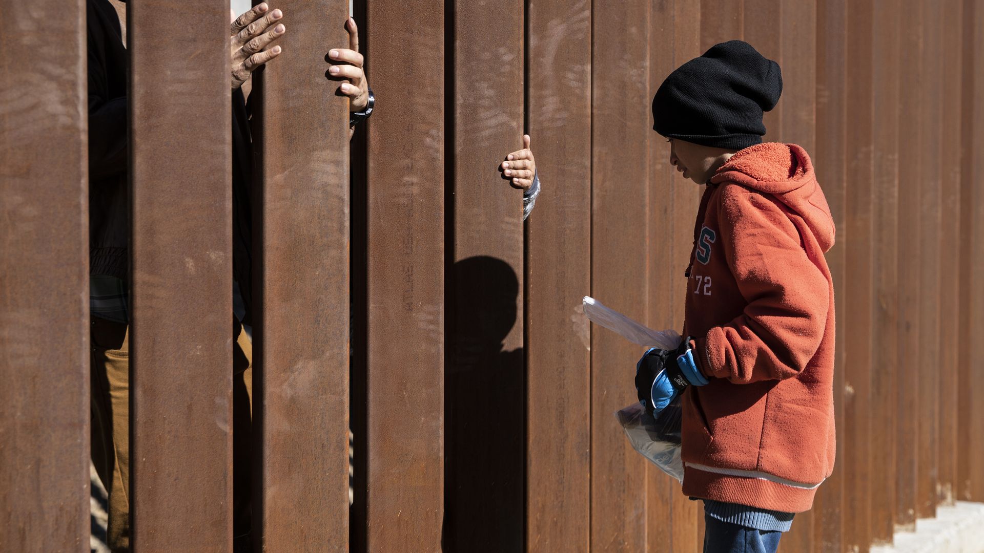 A young migrant boy with an orange hoodie and black hat stands on one side of the border wall.