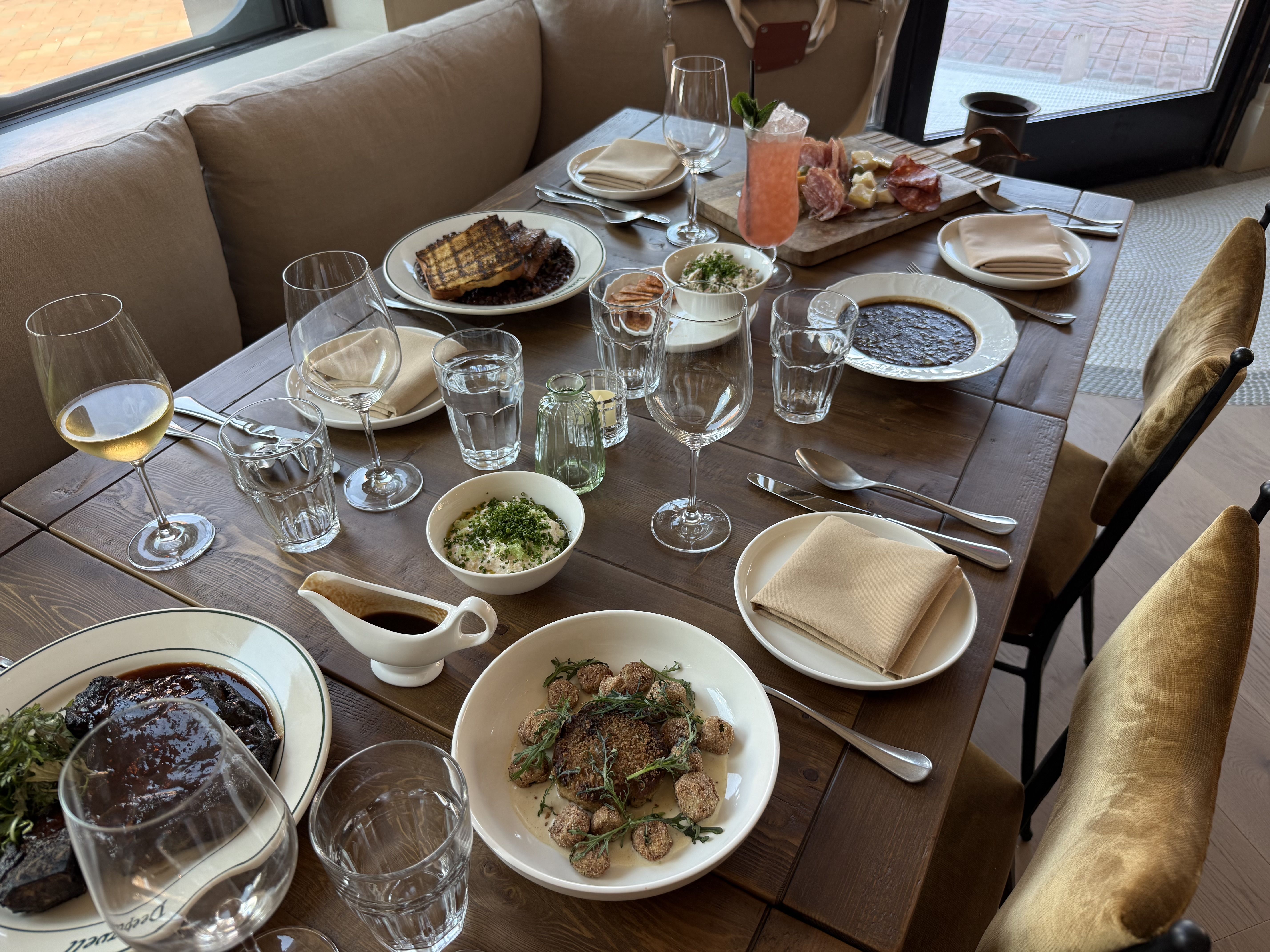 Overhead view of a wooden dining table set for a feast: roasted meat, croquettes, greens, a charcuterie board, bread, wine and a pink cocktail; beige seating and windows in the background.