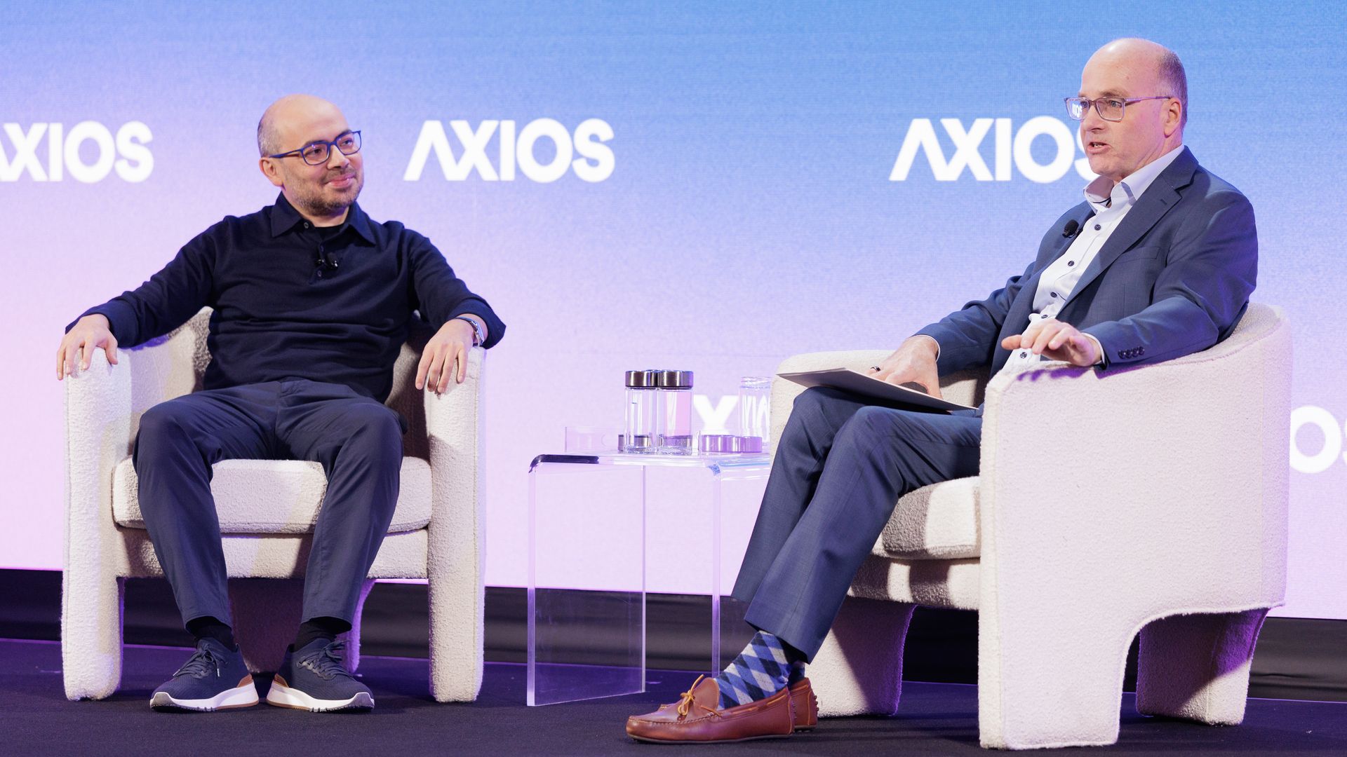 Two men seated in white armchairs on a stage with an "AXIOS" logo backdrop. One in a black shirt and glasses, the other in a blue suit and patterned socks, engaged in conversation.