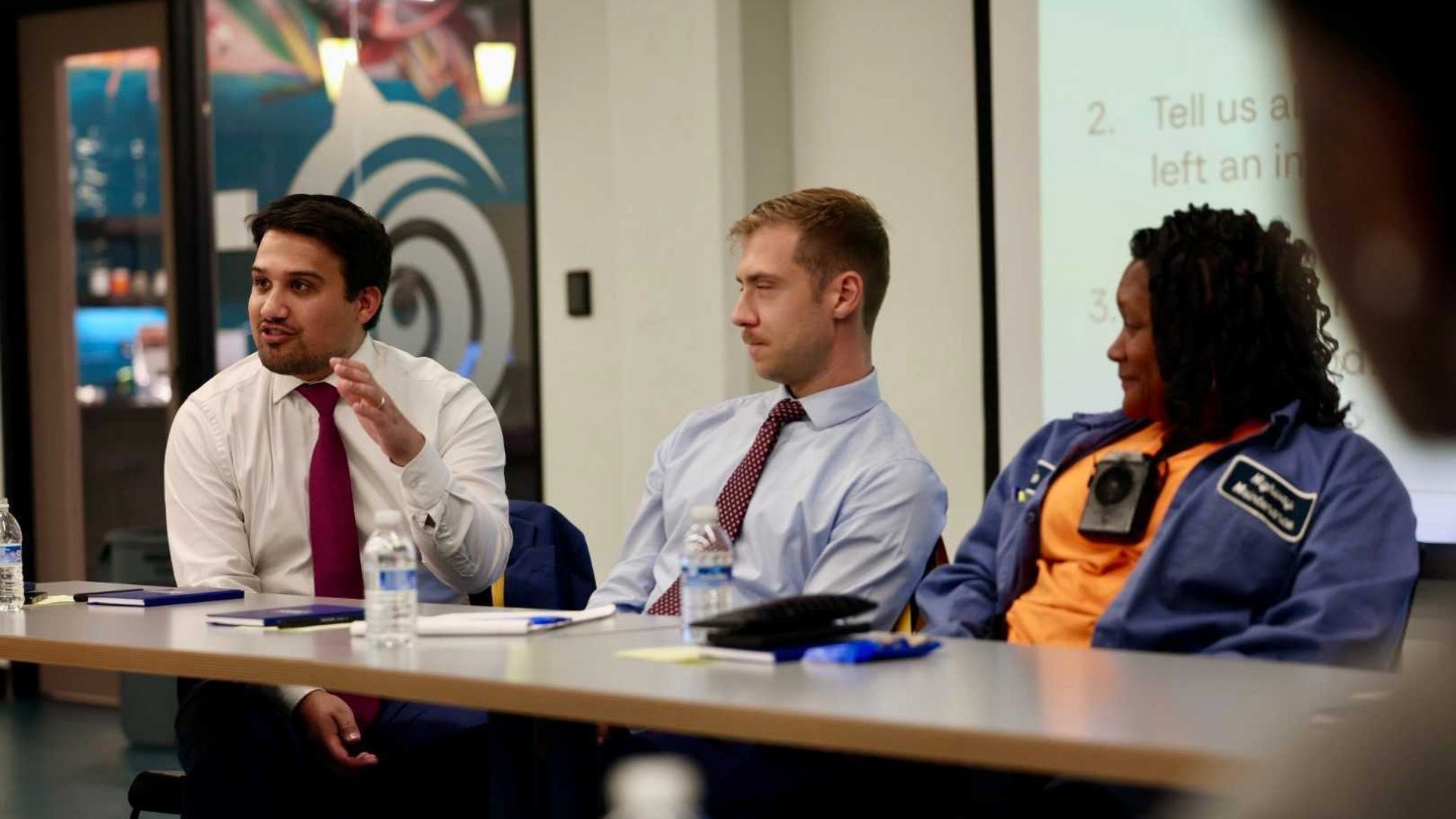 Three people sitting at a table in a meeting room, one man in white shirt and red tie is speaking, another man in light blue shirt and patterned tie listens, a woman in blue jacket and orange shirt looks on.
