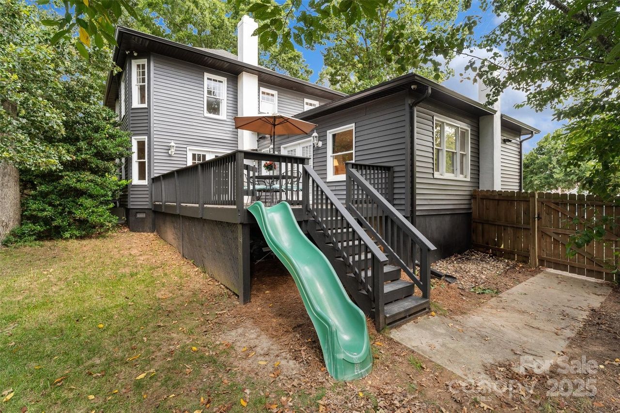 Gray two-story house with white trim, wooden deck with patio table and umbrella, attached green slide, stairs, fenced backyard, surrounded by trees under blue sky.