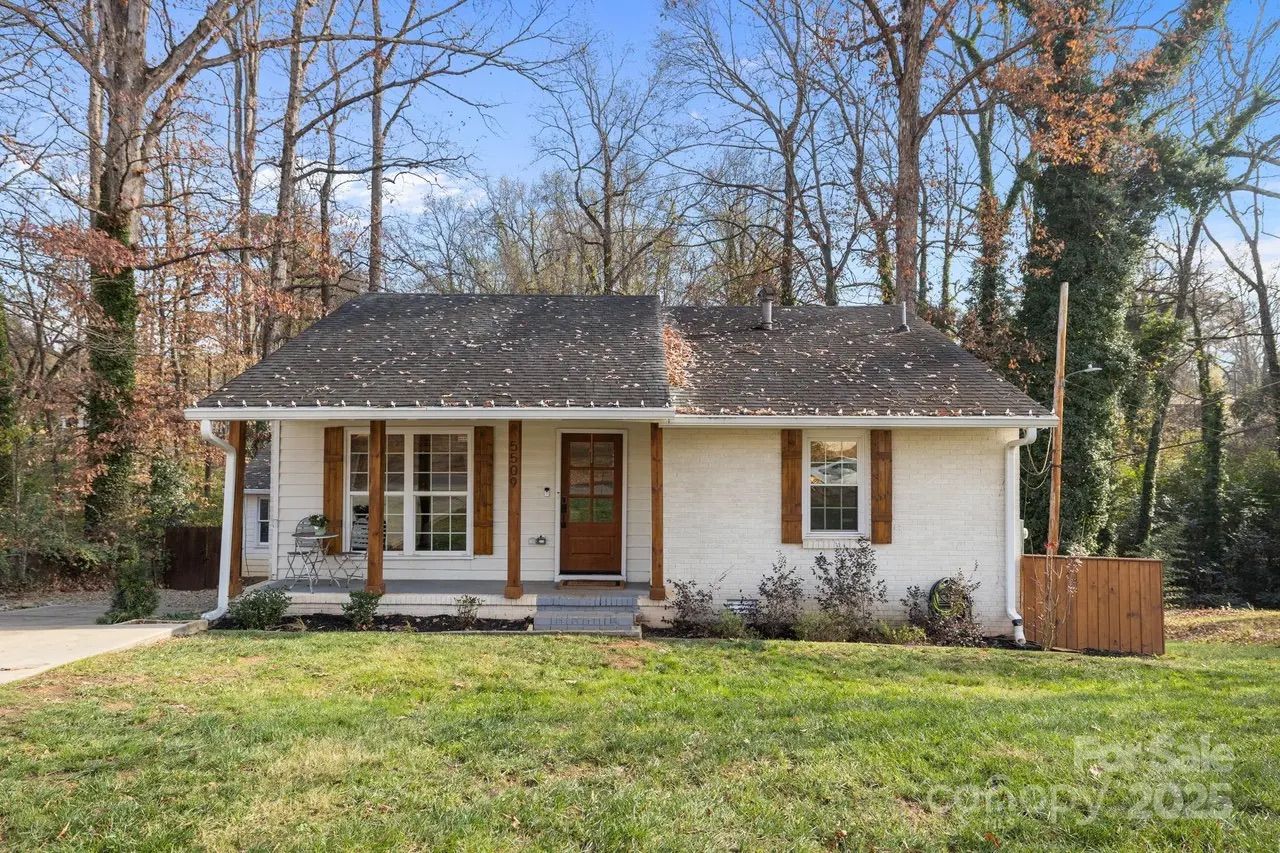 Single-story white brick house with dark shingled roof, wooden door, and windows with wooden shutters, surrounded by leafless trees and green lawn under a clear blue sky.