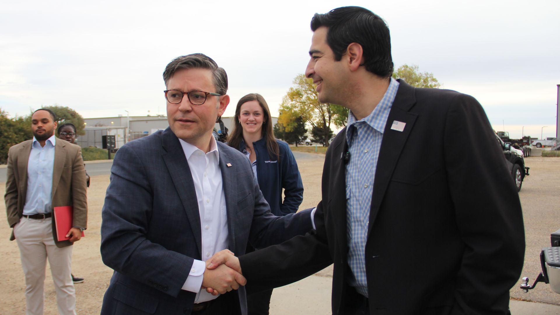 Outside in a parking lot, professionals chat as two men shake hands in the foreground: one in a navy suit with glasses, the other in a dark suit and blue shirt; others stand nearby.