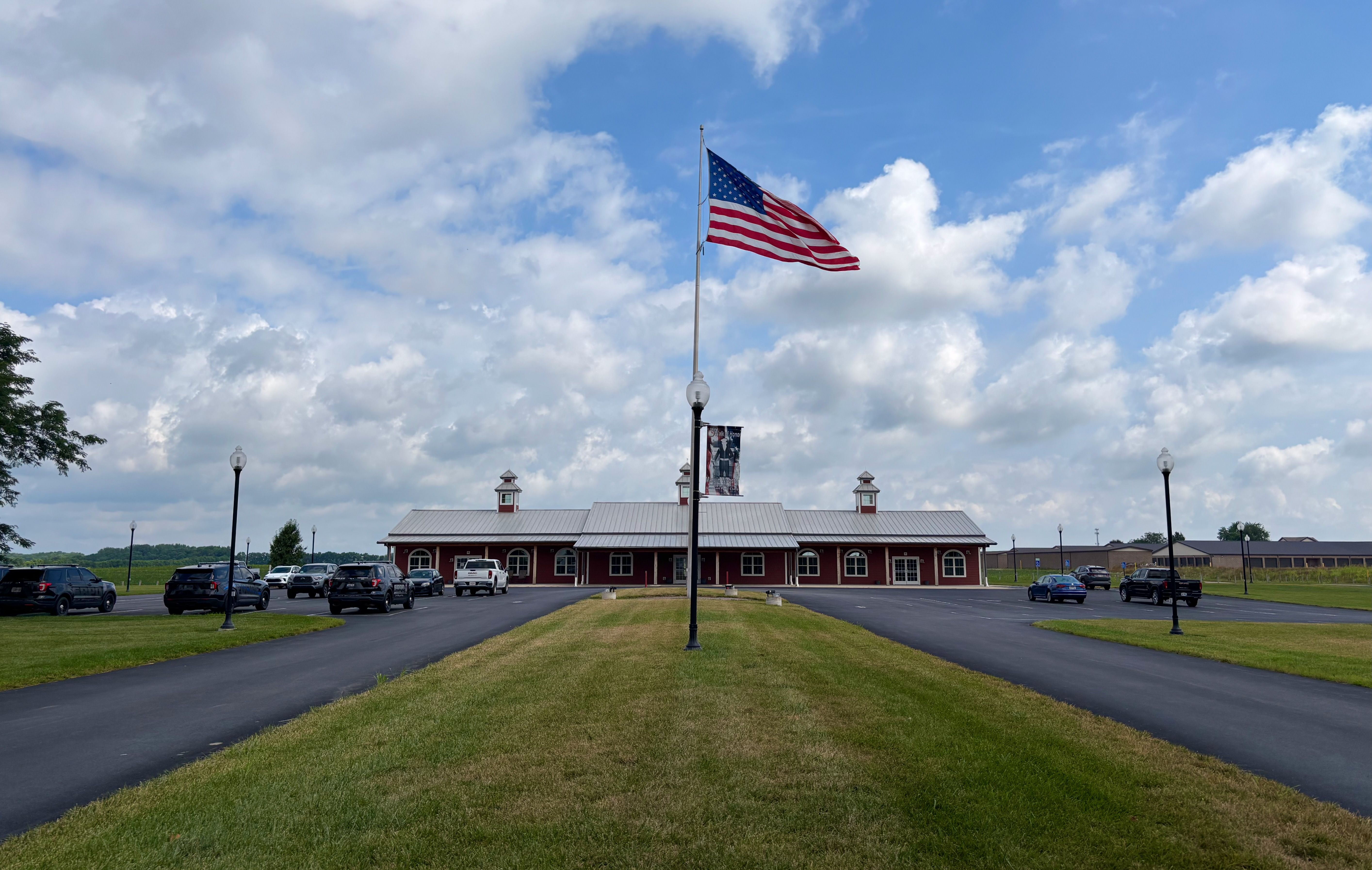 American flag waves atop a tall flagpole in a parking lot in front of a red brick building with a metal roof, under a blue sky with white clouds and several parked cars.