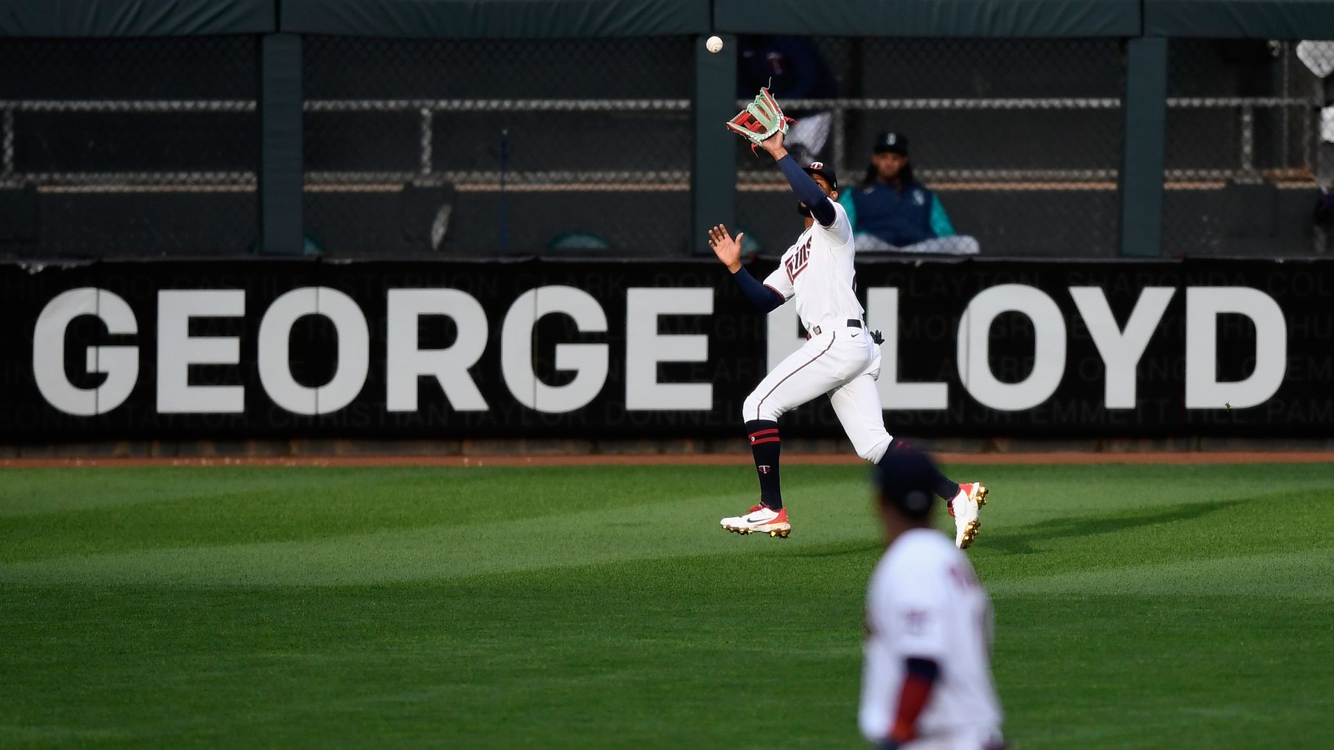 Photo of a baseball player running to catch a ball in a stadium with George Floyd's name hanging as a banner