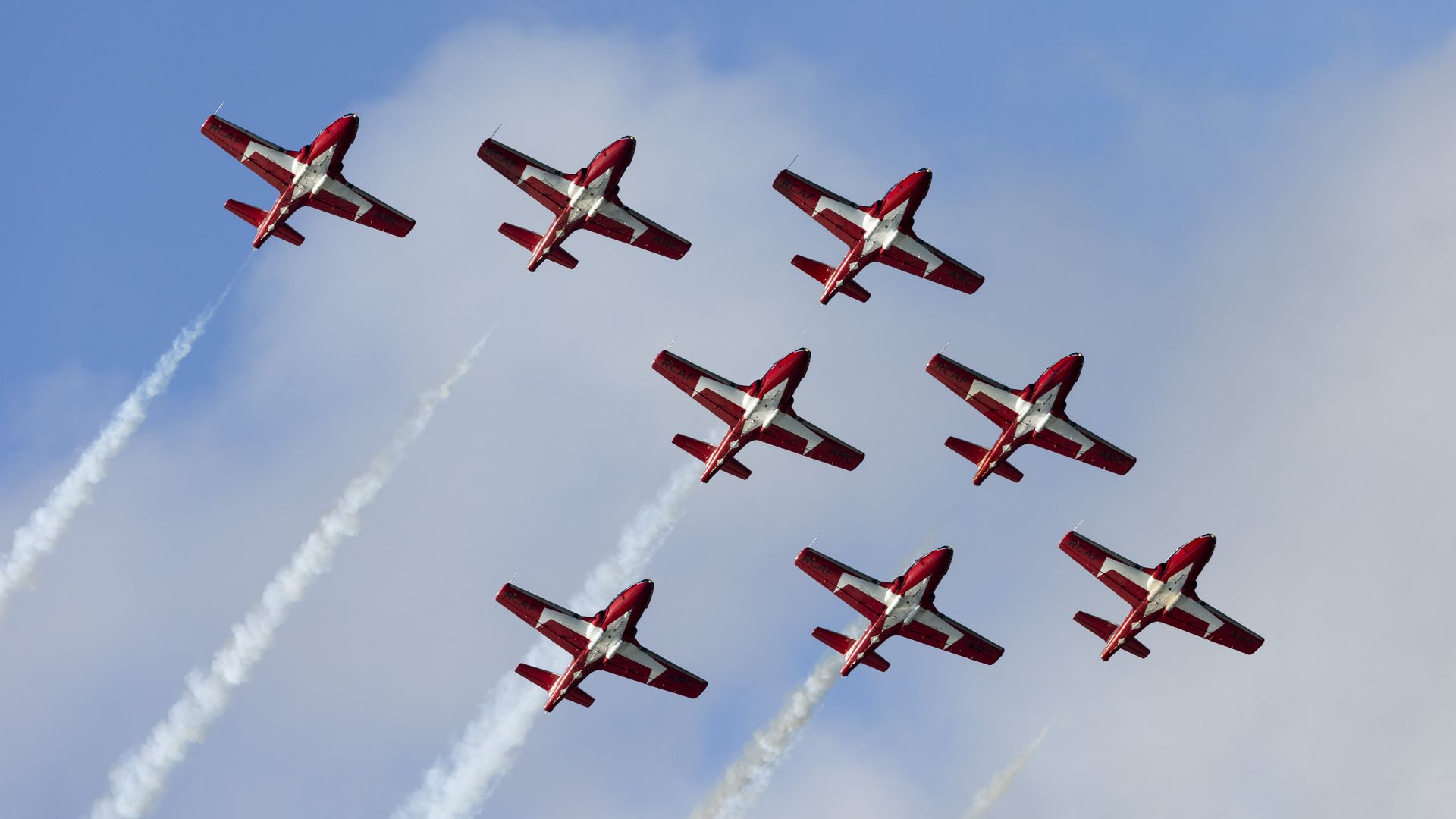 Six red and white aerobatic planes form a diagonal line across a blue sky, each leaving white smoke trails as they perform an air show maneuver.