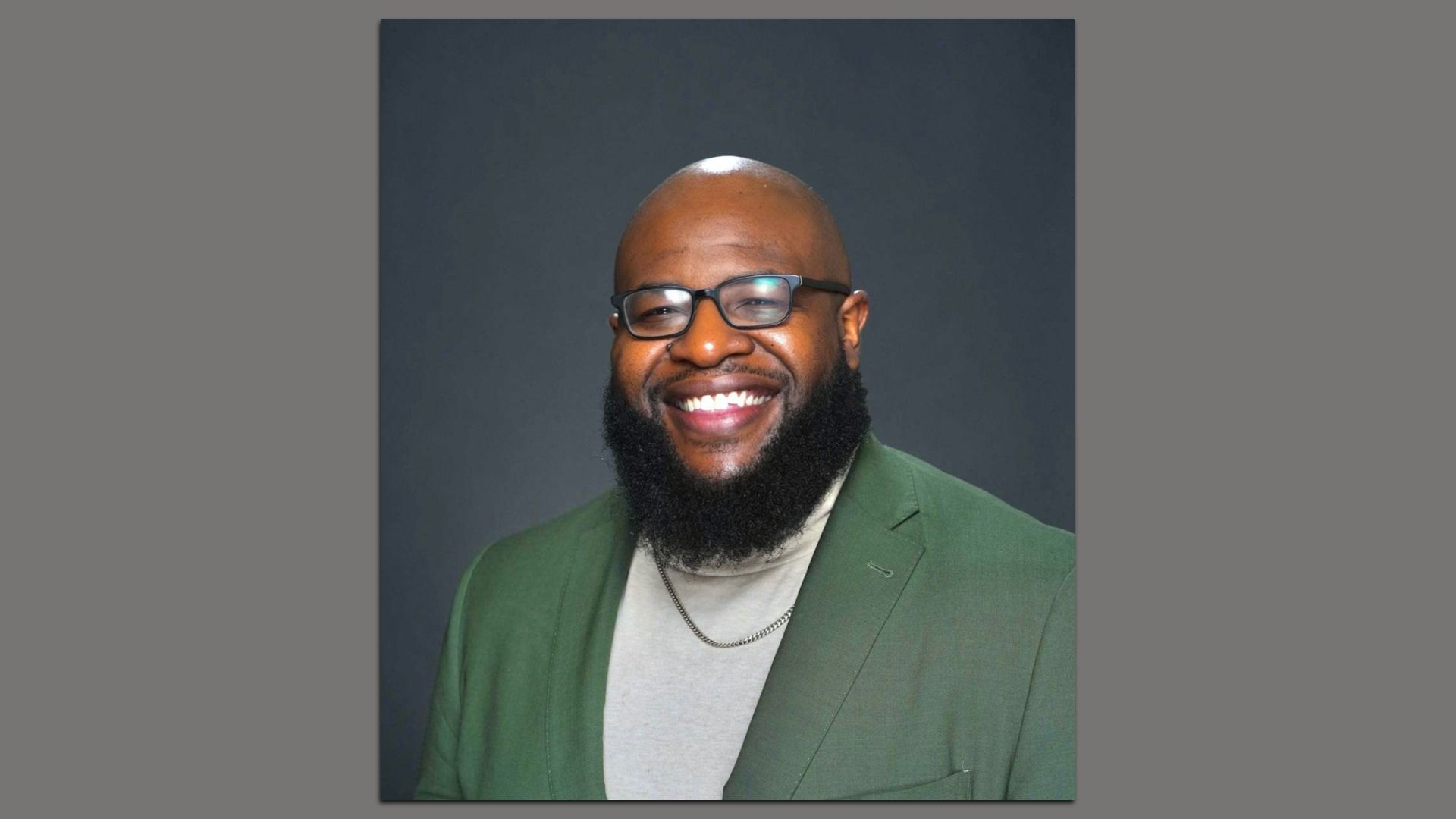 Portrait of a smiling African American man with a full beard and glasses, wearing a green blazer over a light gray shirt, against a dark studio backdrop.