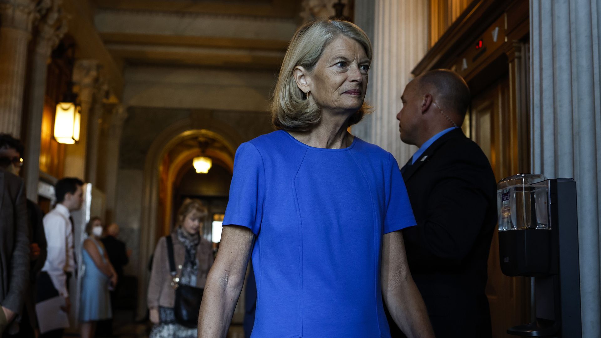 Sen. Lisa Murkowski, wearing a blue dress, walks past a Capitol Police officer into a Republican lunch at the U.S. Capitol.