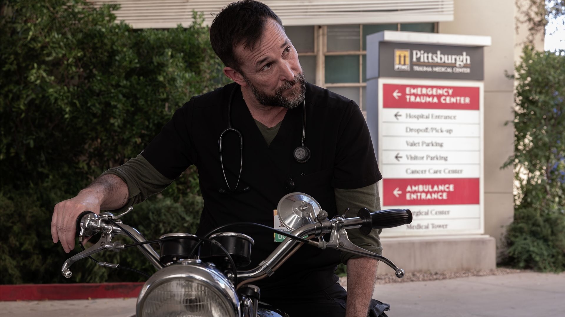 Bearded man in black medical scrubs with a stethoscope sits on a motorcycle outside a hospital; a Pittsburgh Emergency/Trauma Center sign is visible in the background.