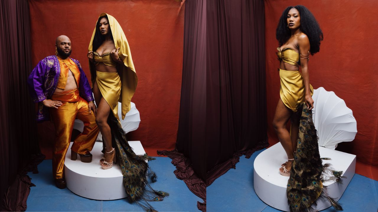 Three models posing on two white circular pedestals against a red backdrop. They wear dramatic translucent ruffle outfits with white headpieces; the woman on the right wears a silver sequin dress.