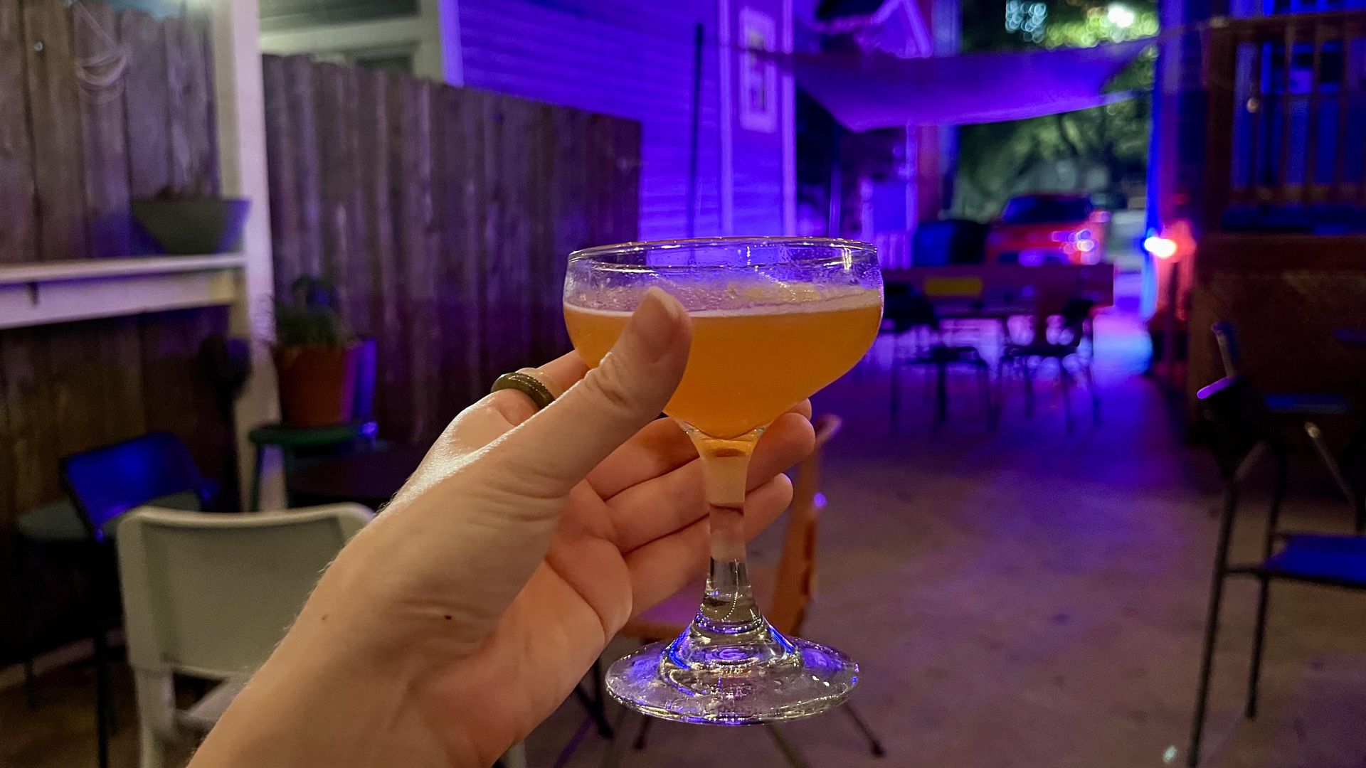 A hand holds an orange-pink colored cocktail in a small coupe glass at Lowcountry. In the background, a backyard patio at night is lit with purple-blue lights and there's a wooden fence and chairs.