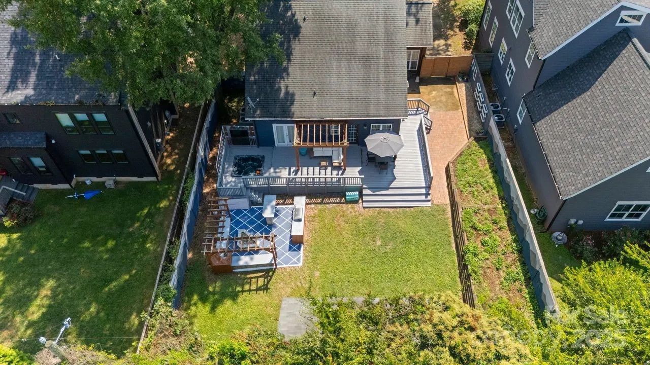 Aerial view of a backyard with a blue-gray single-story house, a deck with pergola and umbrella, outdoor seating area on a blue rug, green lawn, and neighboring houses with fences.