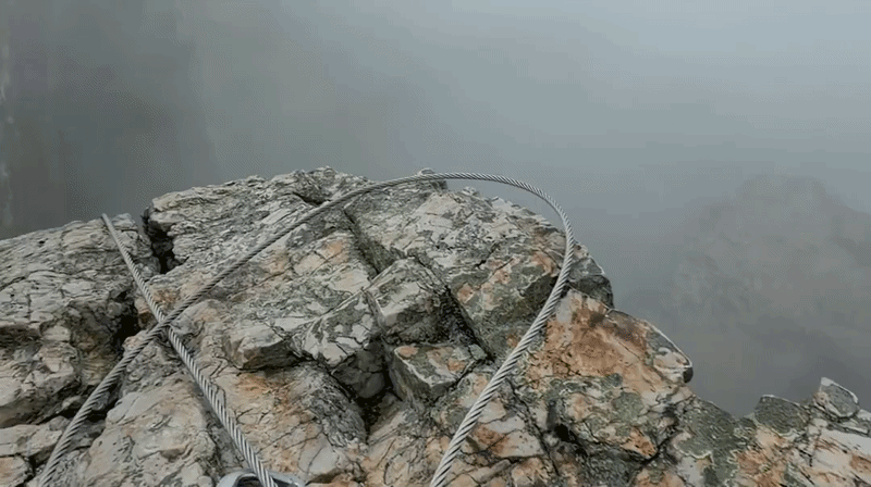 Jagged gray rocks form a cliff edge with a steel cable railing looping across; a misty blue-gray background obscures distant mountains, creating a precarious overlook.
