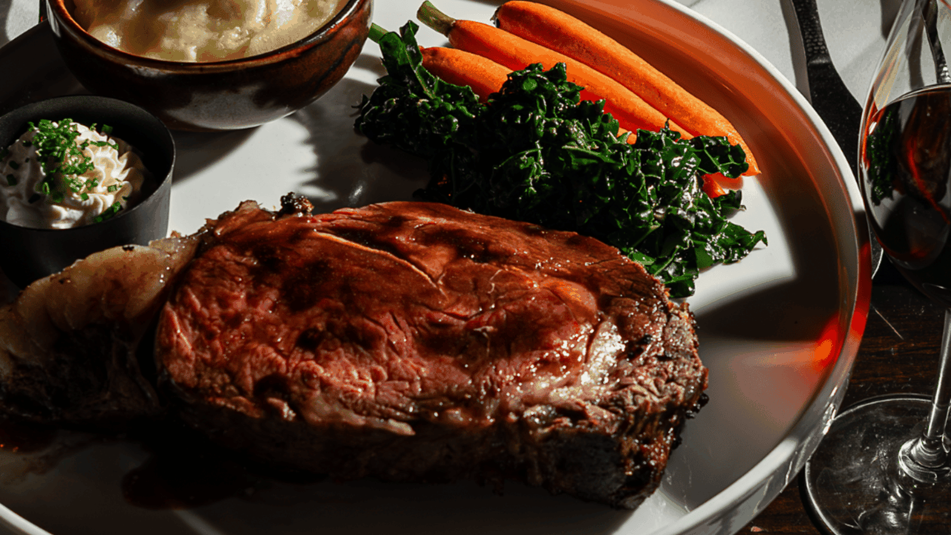 Plate with medium-rare grilled steak, cooked carrots, sautéed greens, and a bowl of mashed potatoes topped with chopped chives, served with a small cup of butter and a glass of red wine on a dark table.