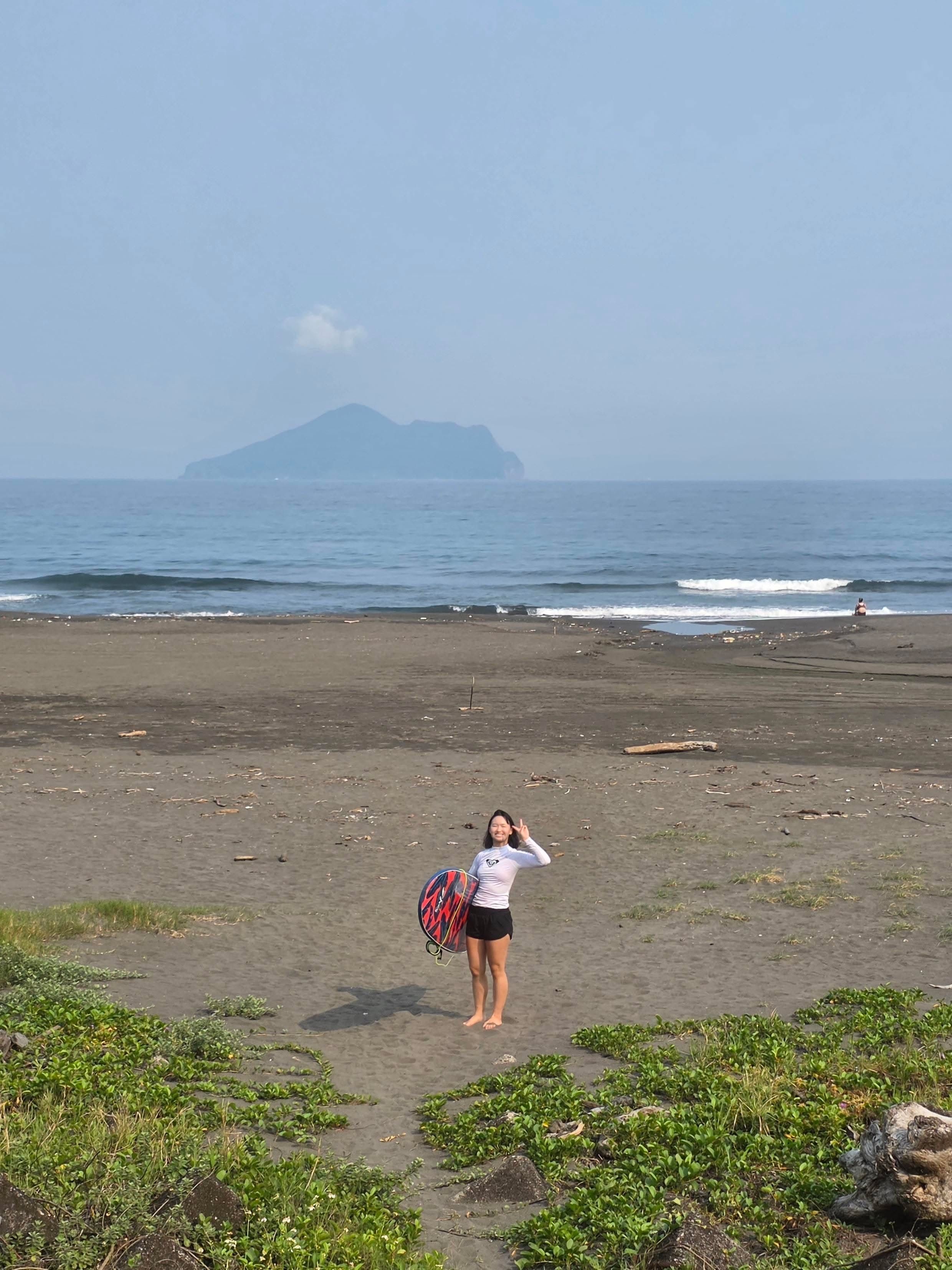 A person on a sandy beach holds a colorful surfboard; calm blue sea with gentle waves and a distant island on the horizon under a pale sky.