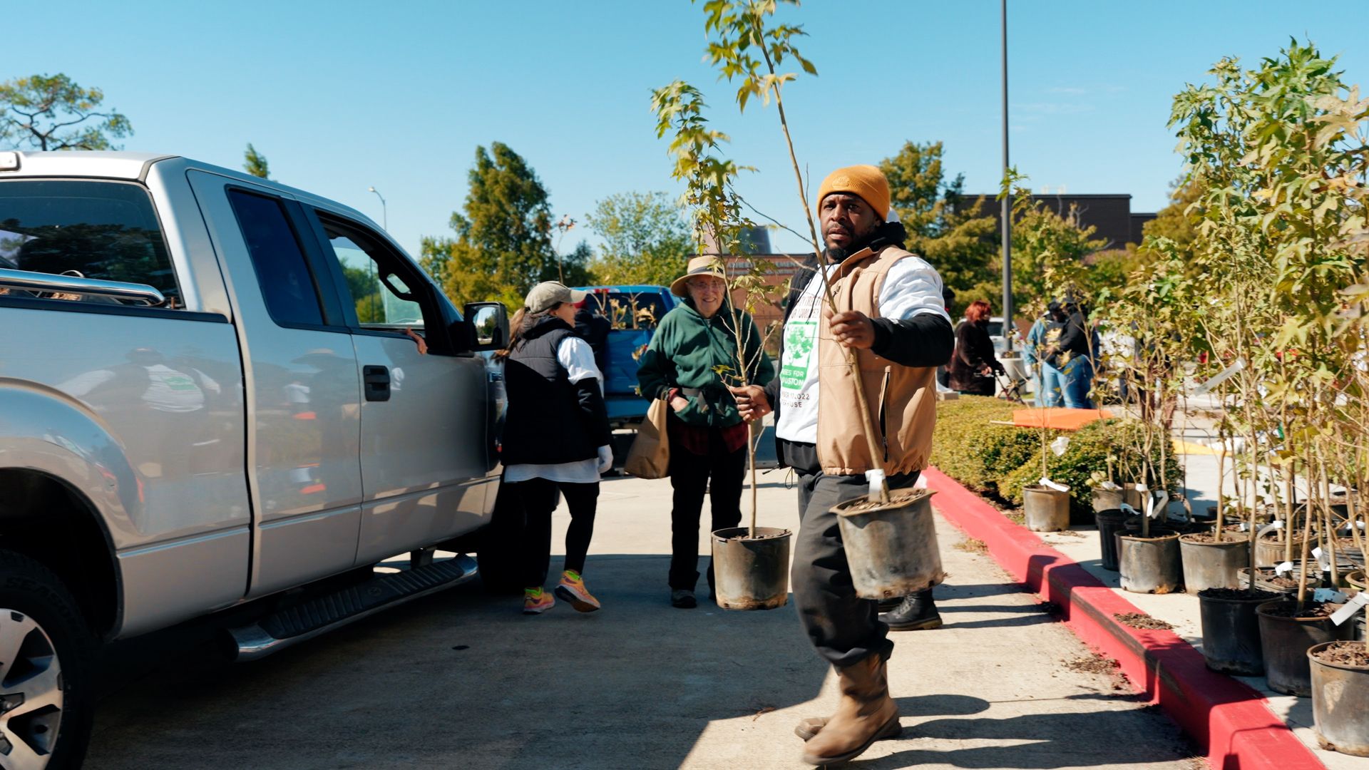 A Trees for Houston volunteer wearing an orange beanie and tan vest carries two saplings 