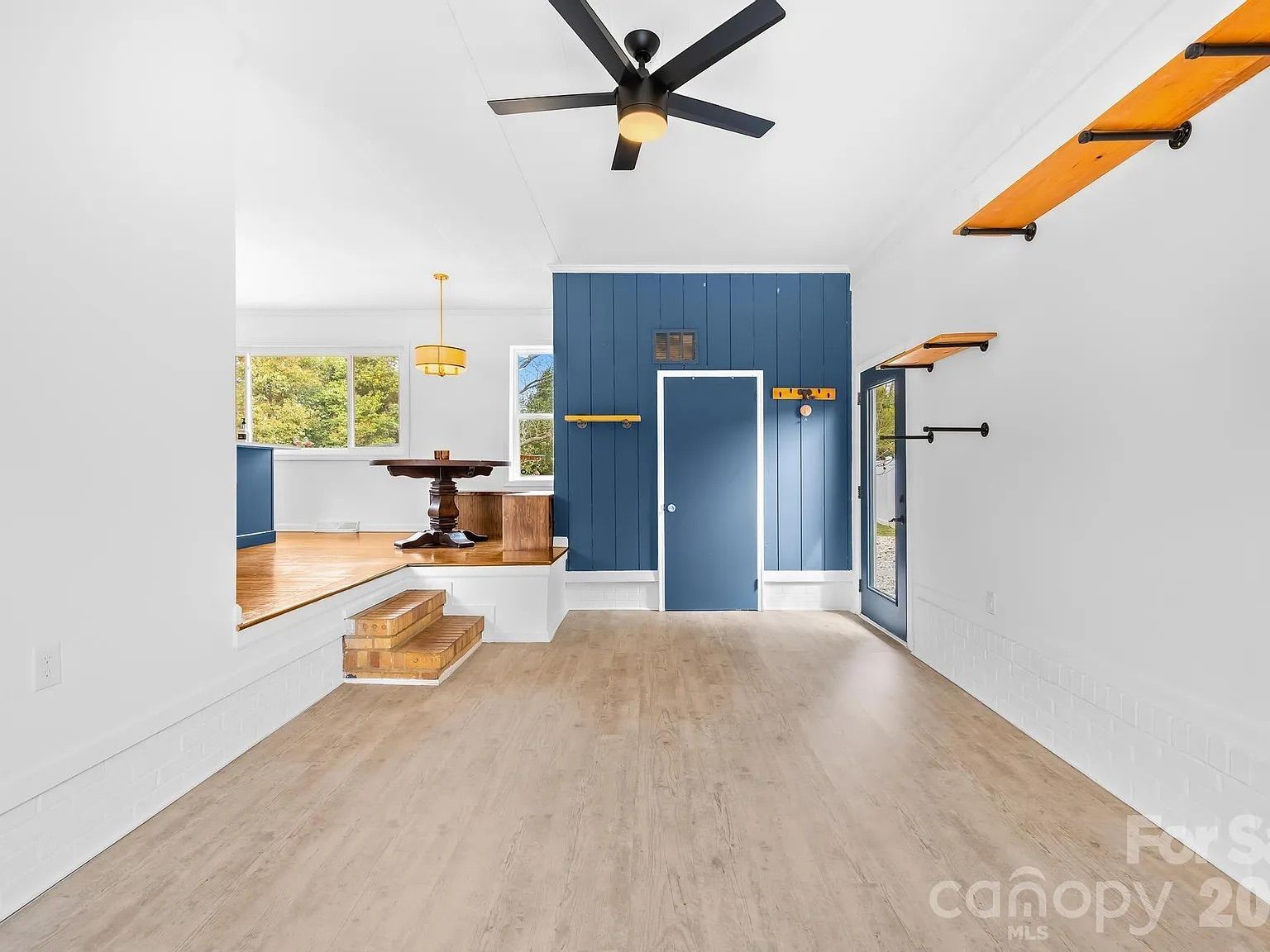 Bright room with light wood floor, white walls, blue door and accent wall, black ceiling fan, wooden shelves, and a wooden table visible on a raised platform with windows showing greenery outside.