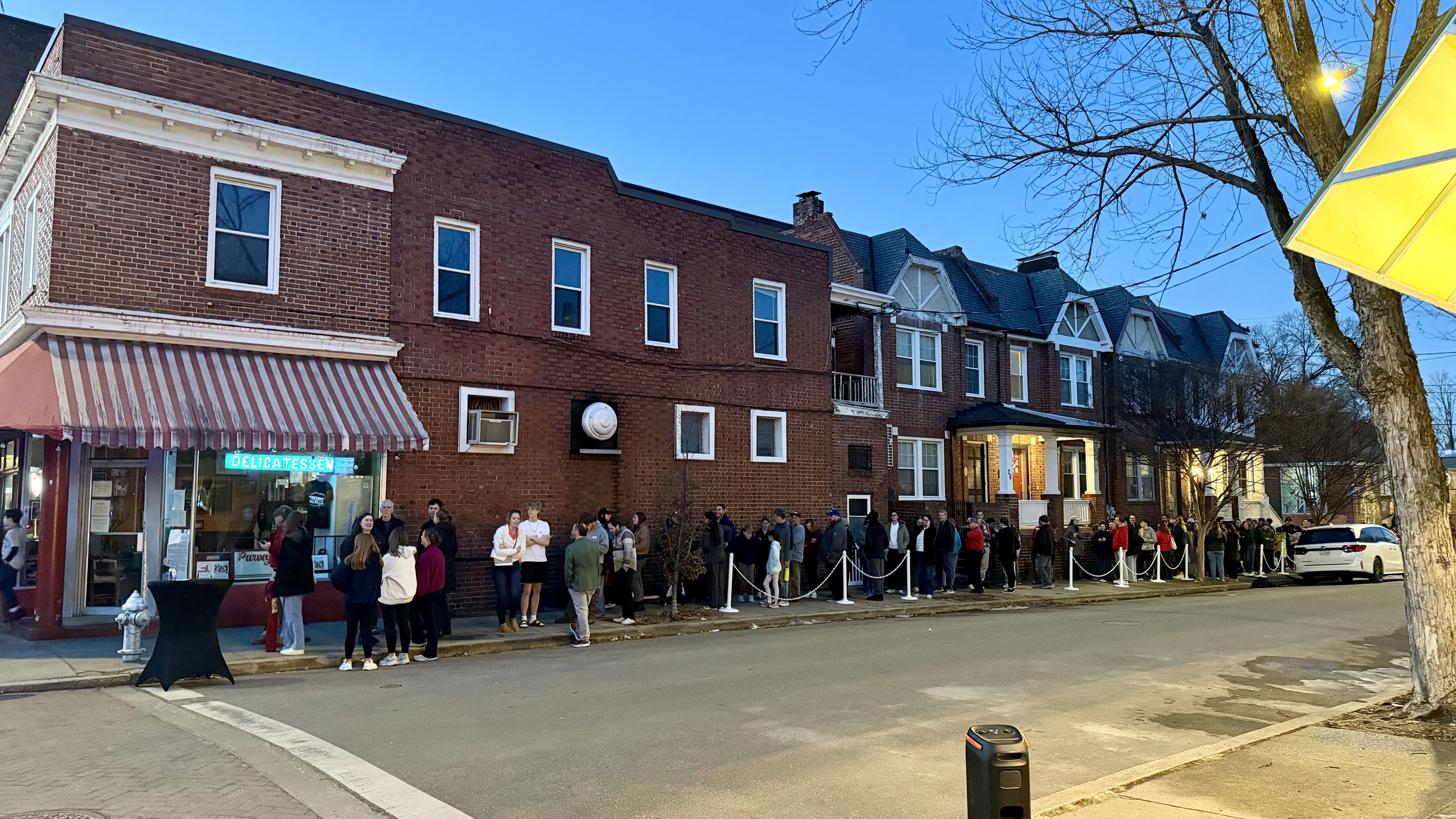 Long line of people outside a brick delicatessen with a red and white striped awning during early evening on a quiet street with residential houses and leafless trees.