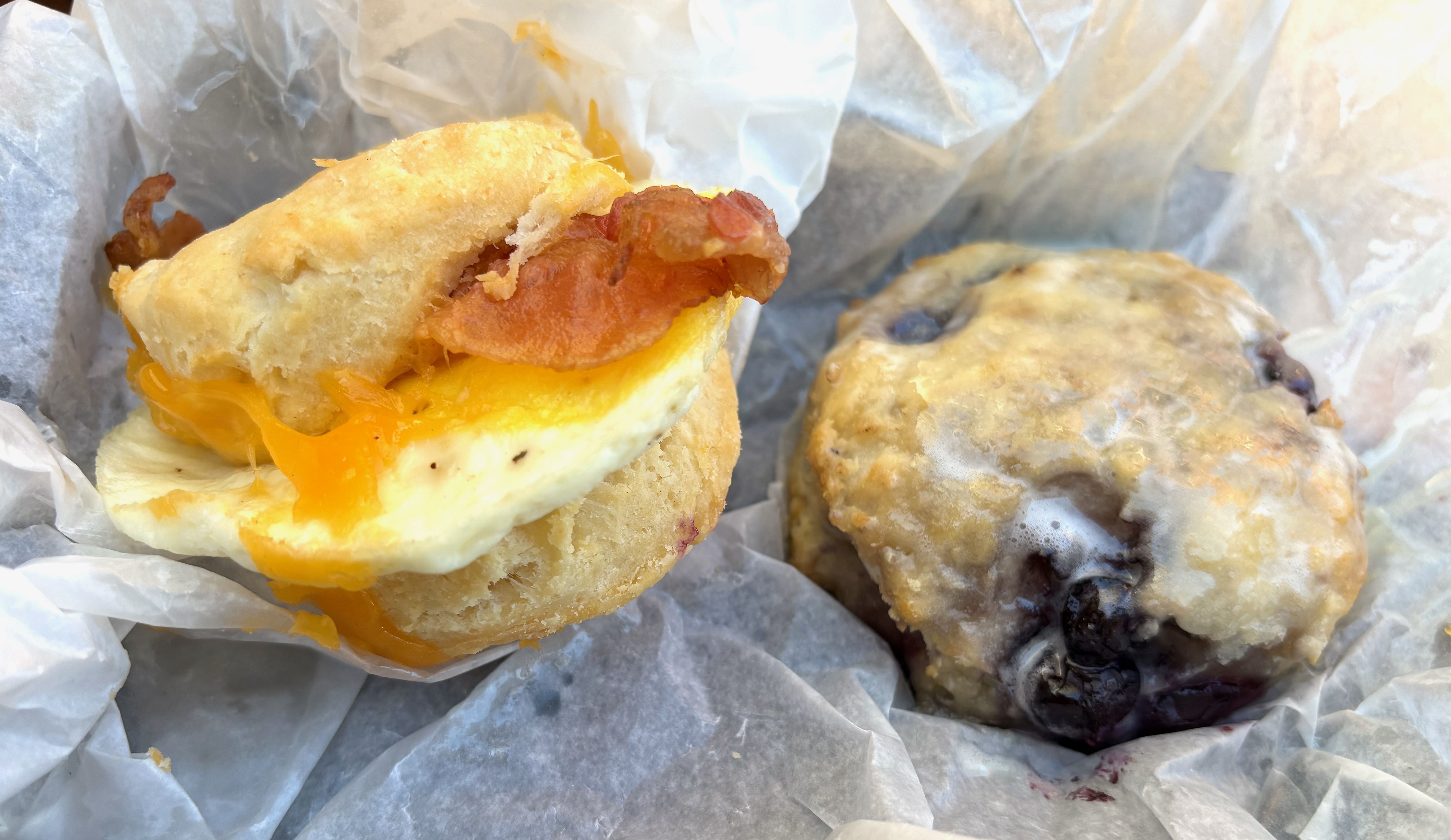 An egg, cheddar cheese and bacon biscuit sandwich and a blueberry biscuit on white food paper