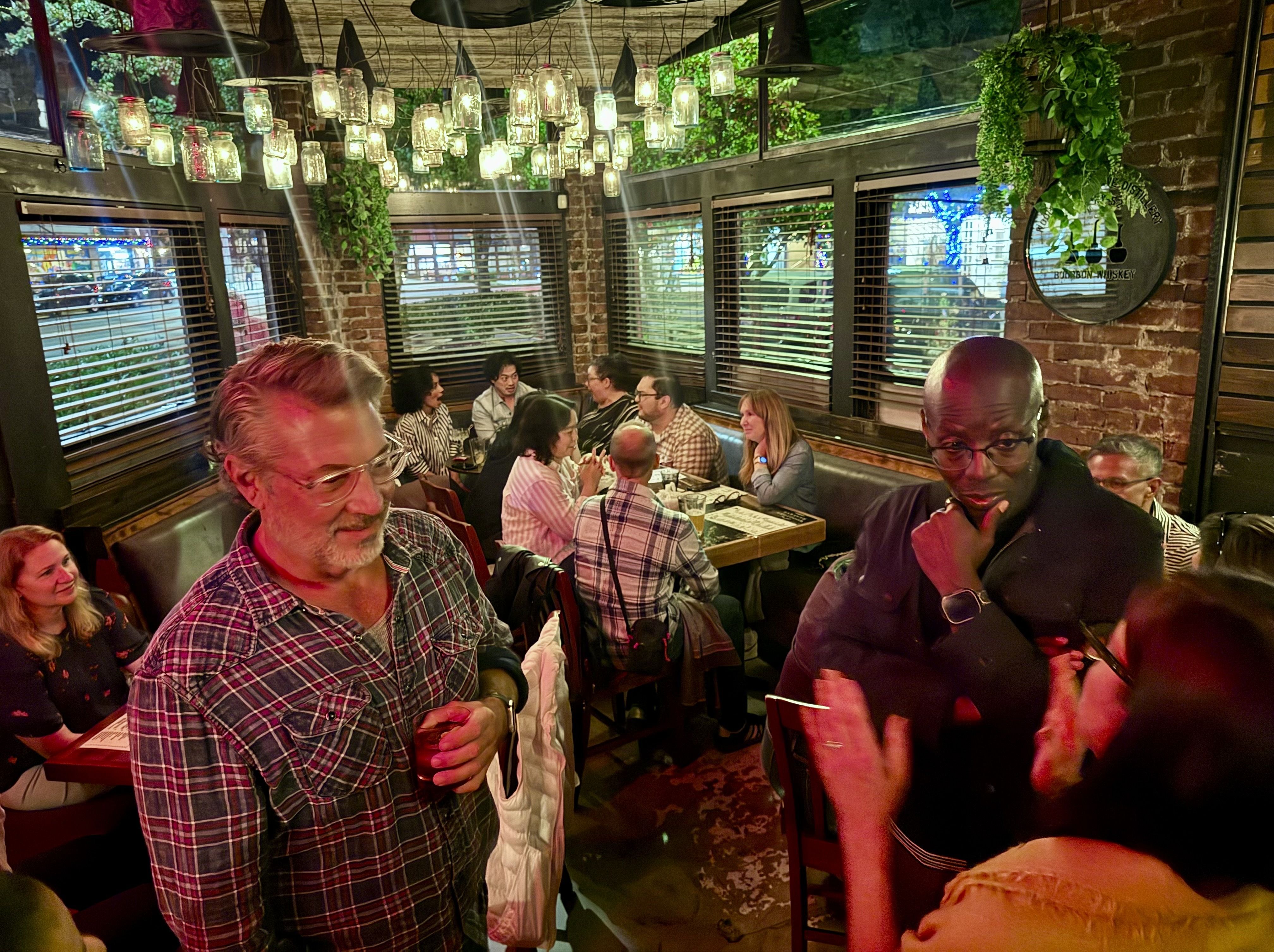 Cozy brick-walled restaurant with hanging mason jar lights and trailing greenery. A lively group of people in plaid shirts sits around a table, chatting and sipping drinks near the windows.