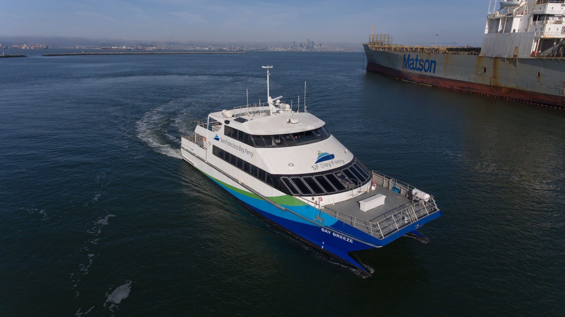 Aerial view of a white SF Bay Ferry called Bay Breeze cutting through blue water toward the right, beside a large Matson cargo ship; distant city skyline on the horizon.