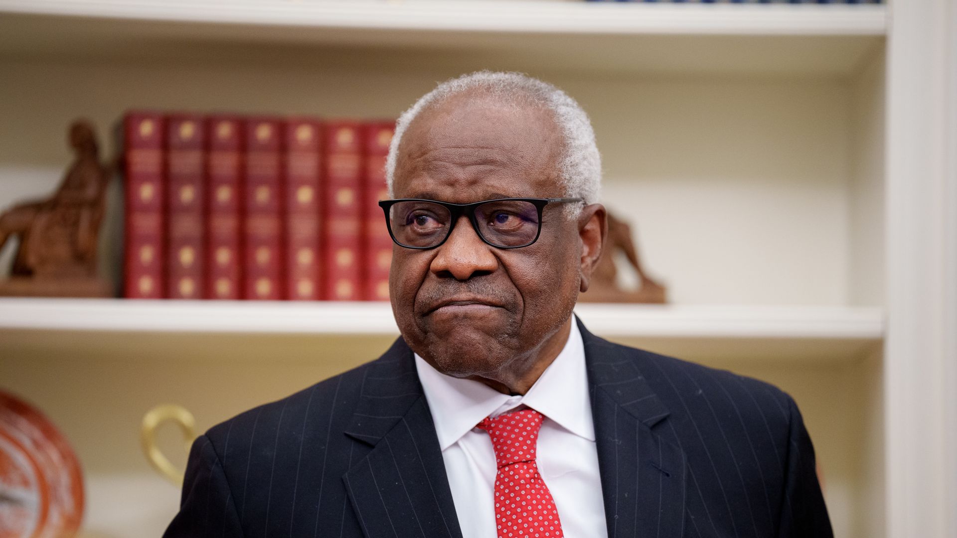 An older man with gray hair and glasses, wearing a dark pinstripe suit, white shirt, and a red polka-dot tie, stands before a bookshelf filled with red books in a study.