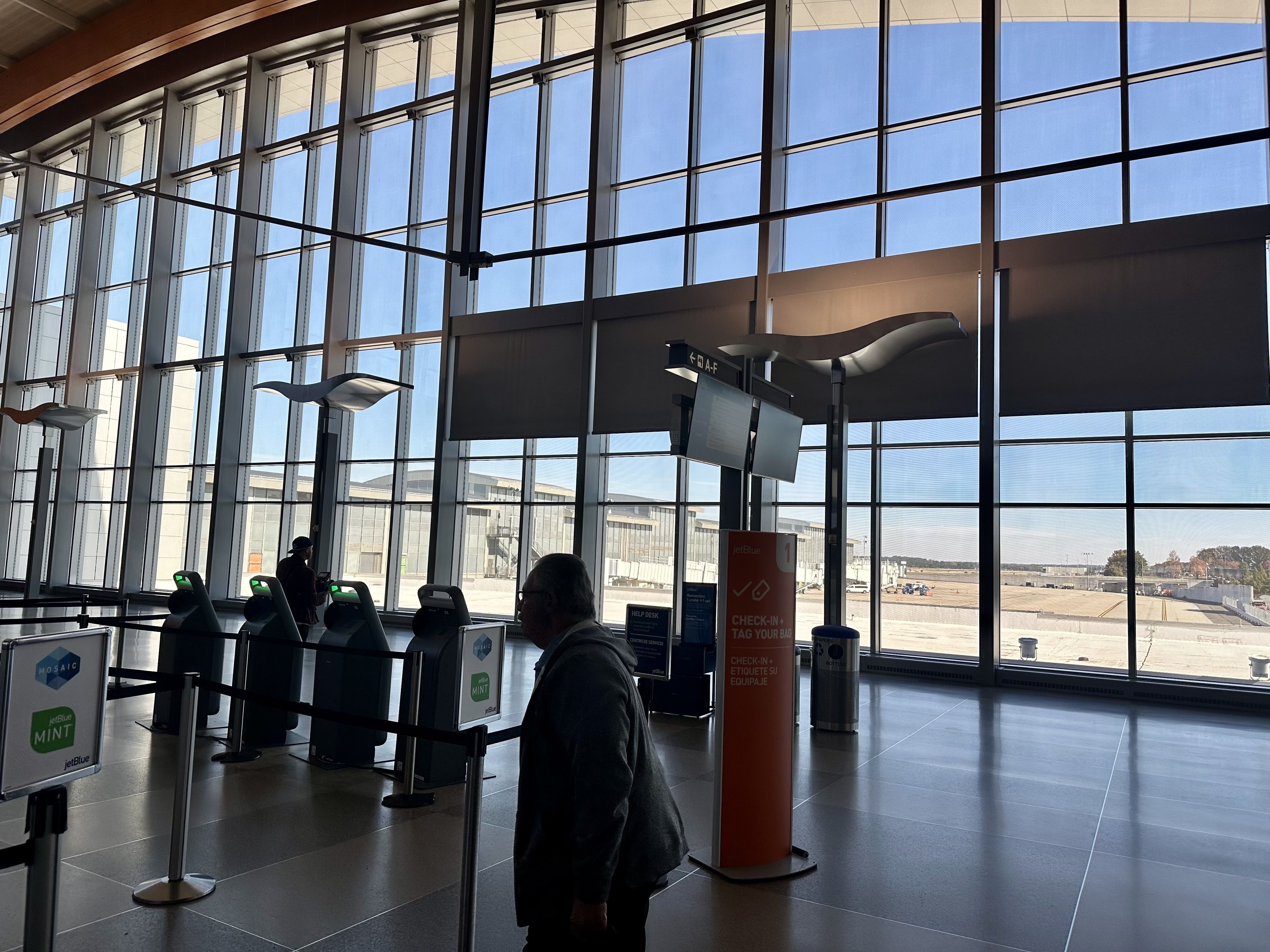 Interior of an airport terminal with large windows showing clear blue sky and runway, several self-service kiosks with JetBlue Mint branding, and a person walking near a JetBlue check-in sign.