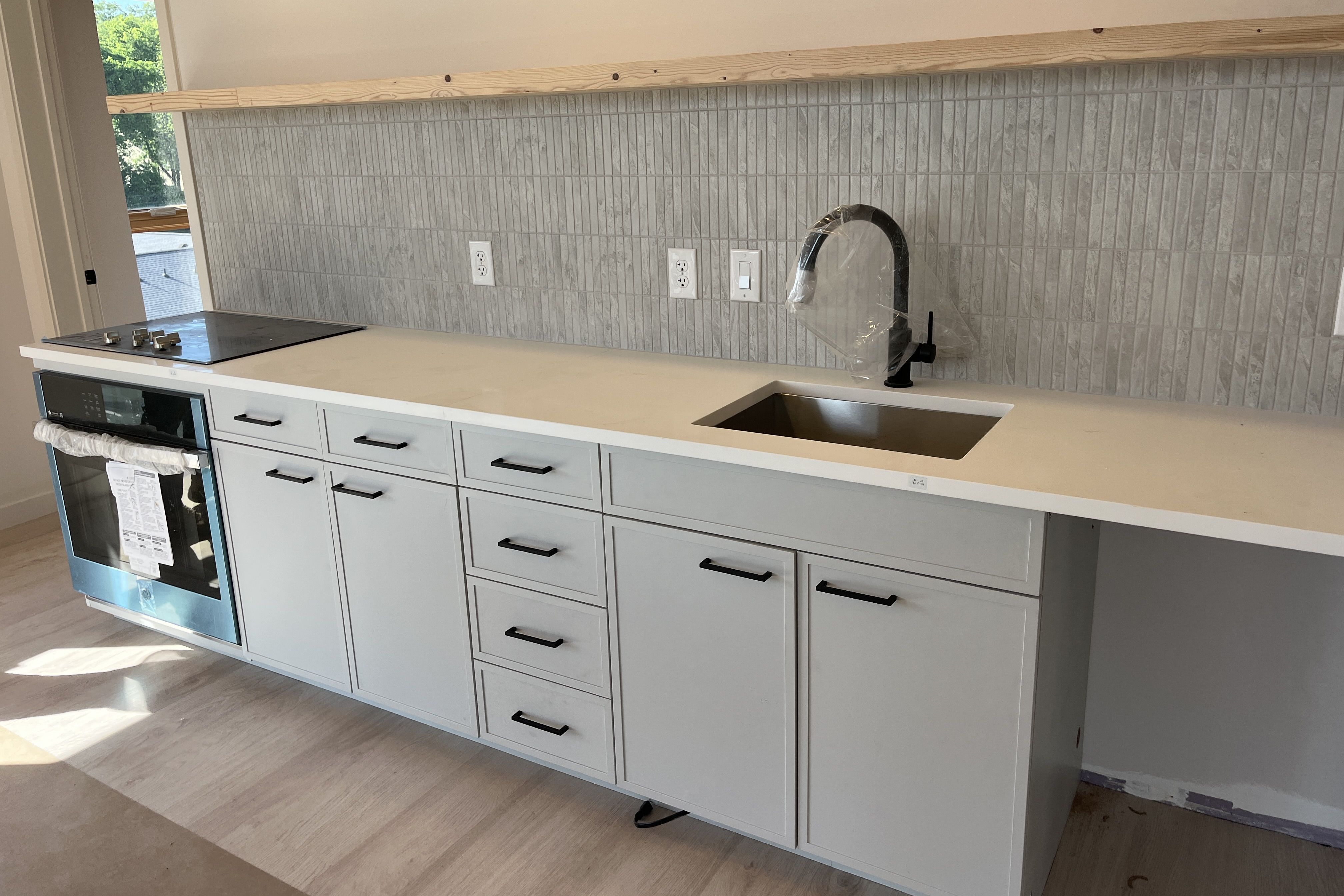 Modern kitchen with gray cabinets, white countertop, and a vertical gray tile backsplash. A black curved faucet over a stainless sink wrapped in plastic. Left side has a stove/oven; natural light from a window.