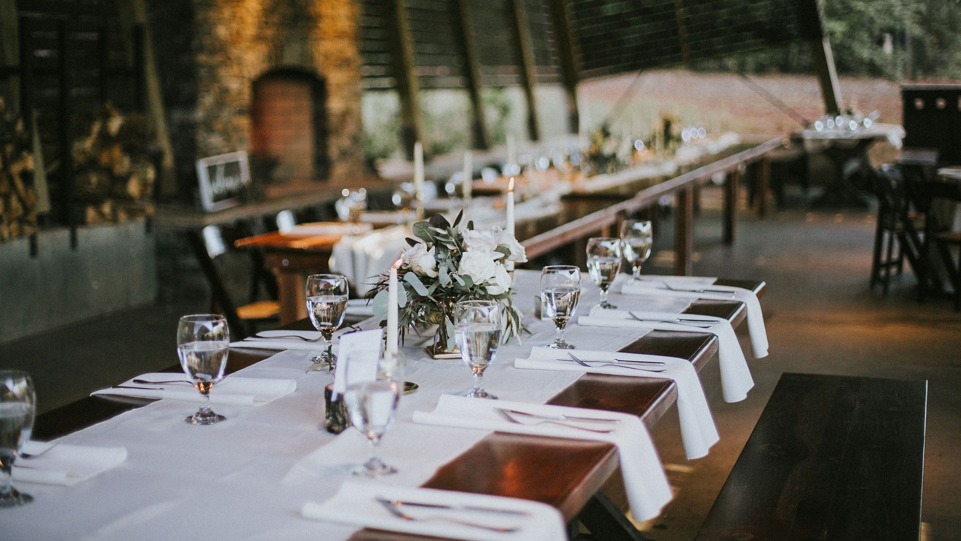 Long wooden table set for a formal event with white table runners, glassware, white candles, and floral centerpieces inside a rustic, dimly lit venue with wood and brick walls at the Whitewater Center. 