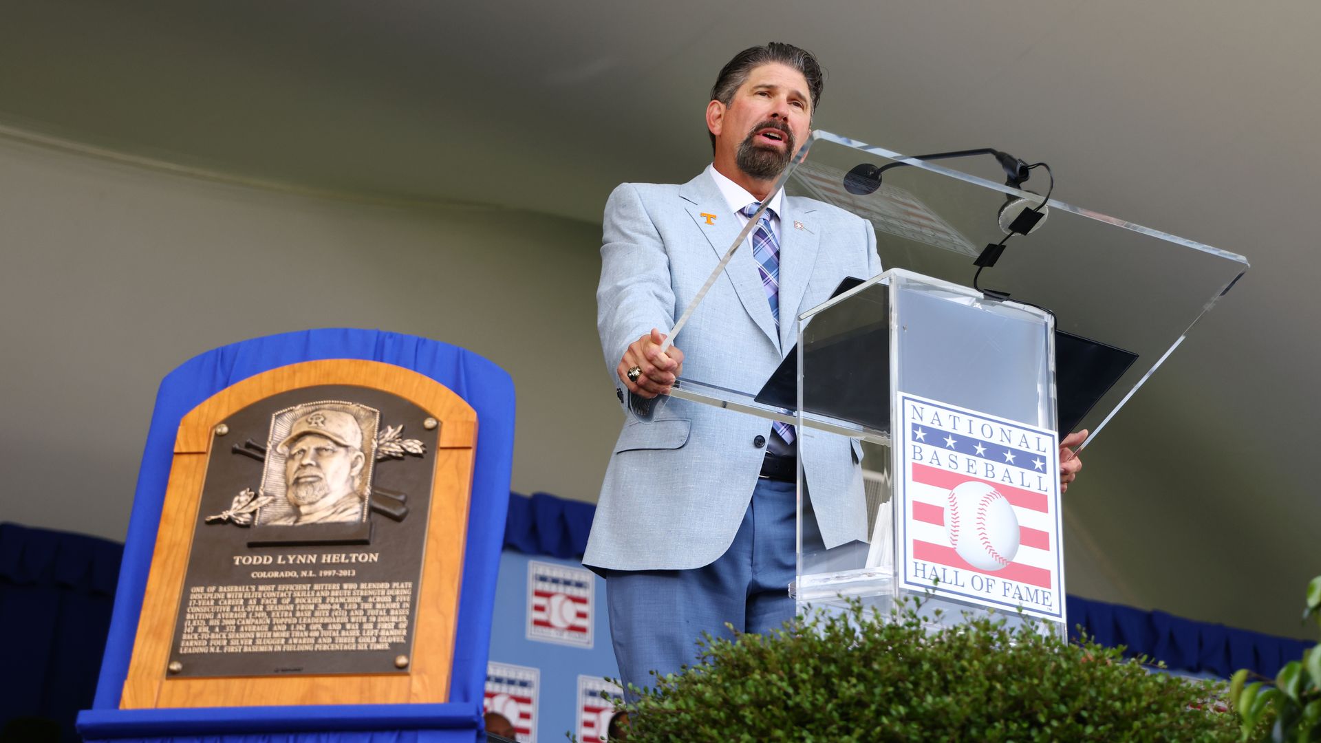 A man in a light blue suit speaks in front of a plastic lectern, with a small plaque depicting his likeness next to him.