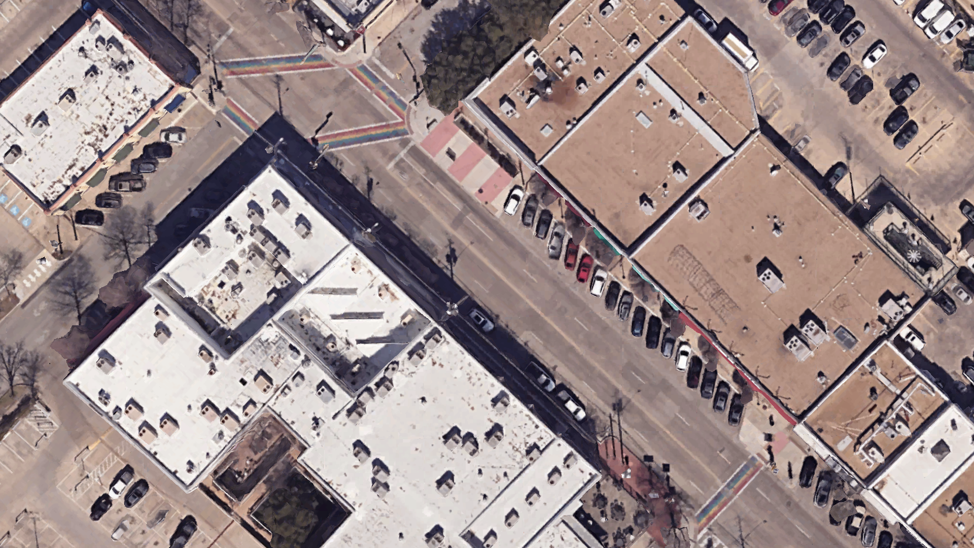 Aerial view of a city intersection with multiple parking lots, several vehicles, and rainbow-colored crosswalks connecting sidewalks around various buildings.