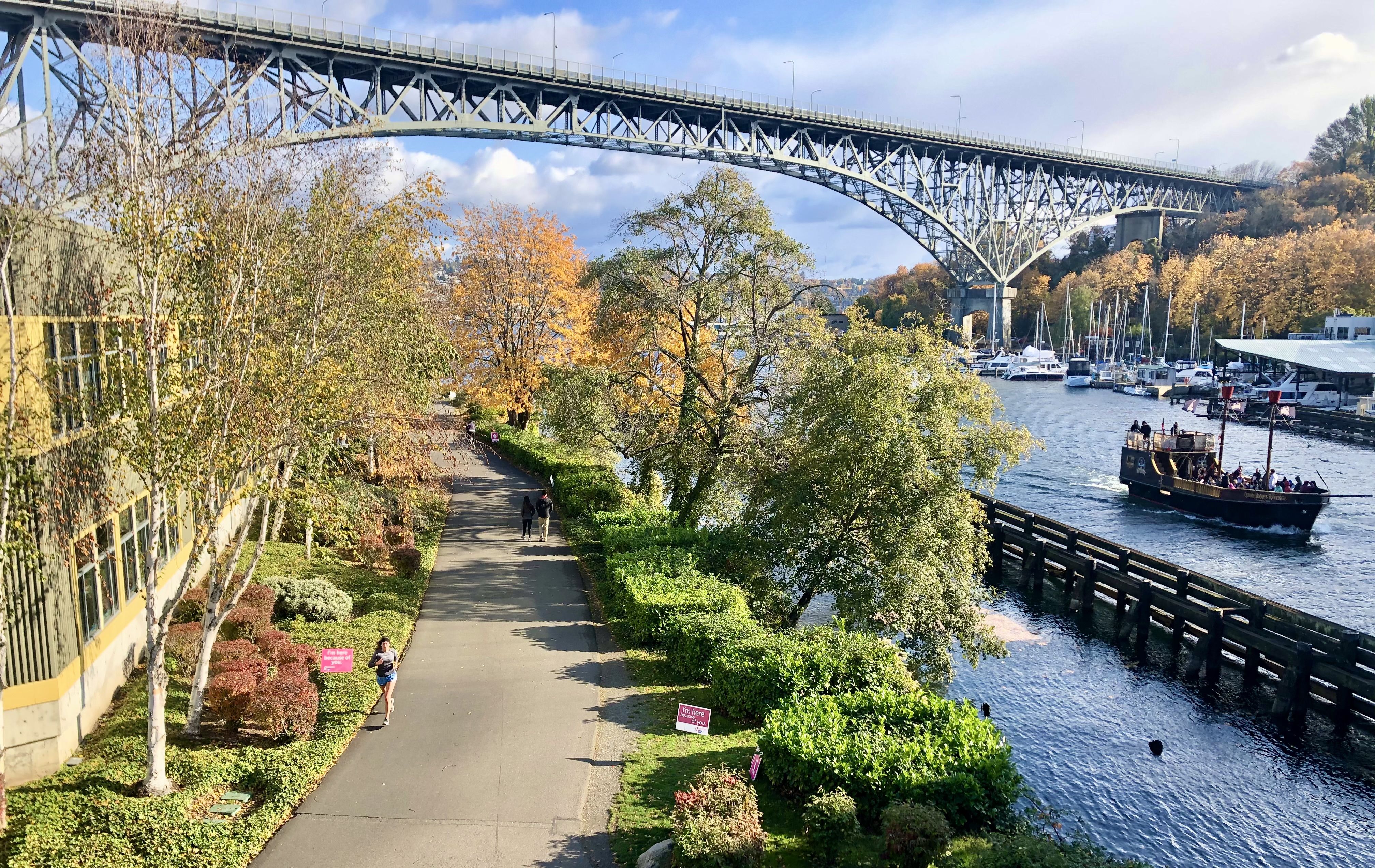 A boat zips by in the Fremont Canal alongside the tree-lined Burke Gilman Trail, with the Aurora Bridge and Lake Union in the background.