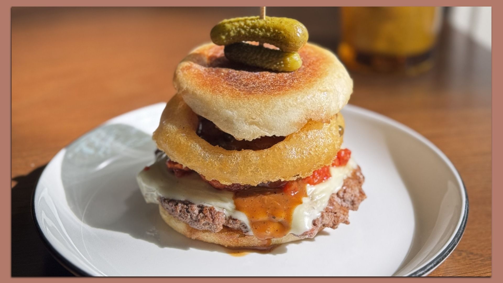 Burger with melted cheese, crispy onion ring, tomato sauce, and two pickles on toasted bun, served on white plate with wooden table background.