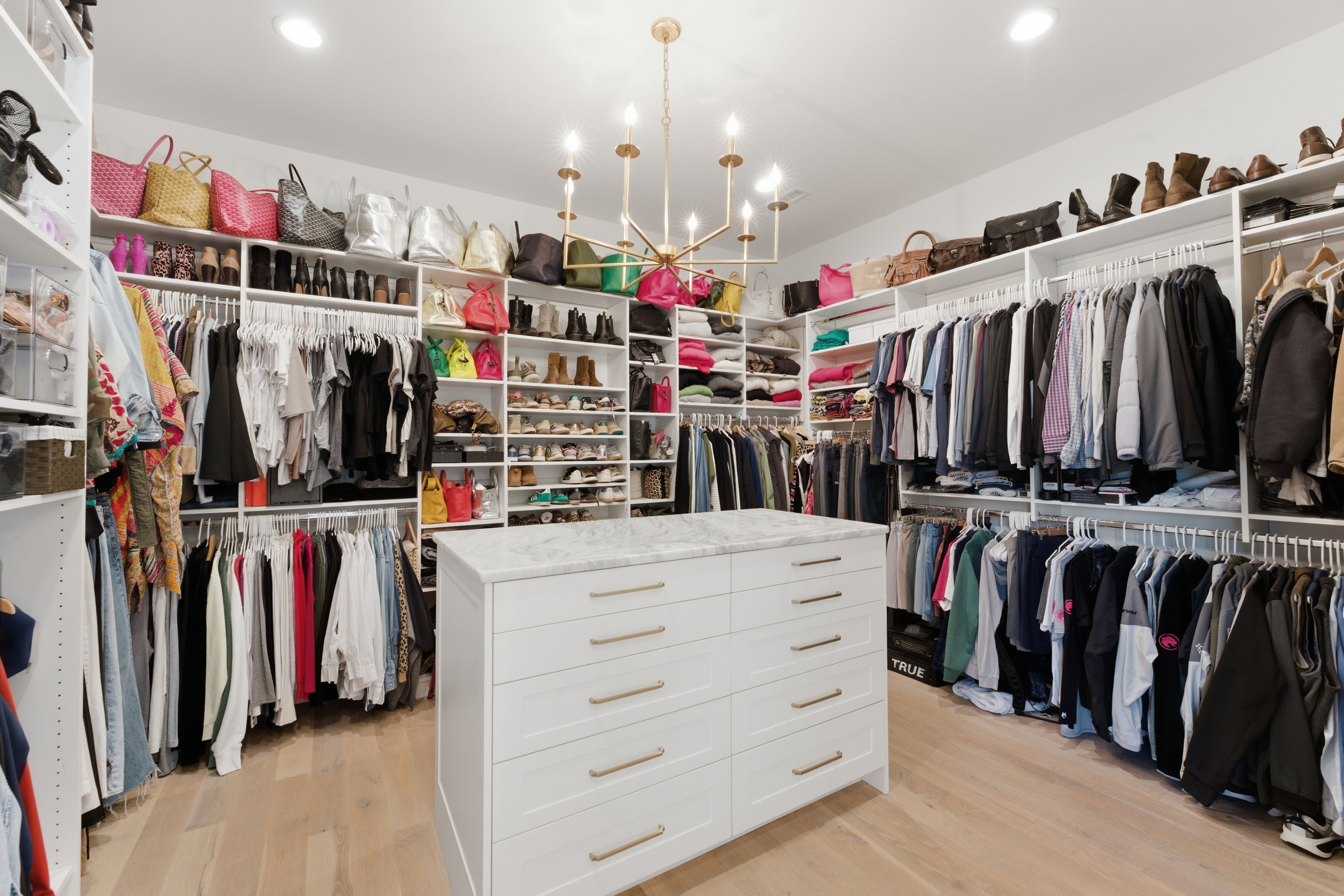A spacious walk-in closet with white, open shelving and hanging racks of clothes, shelves full of shoes and handbags, a marble-topped white island in the center, and a gold chandelier overhead.