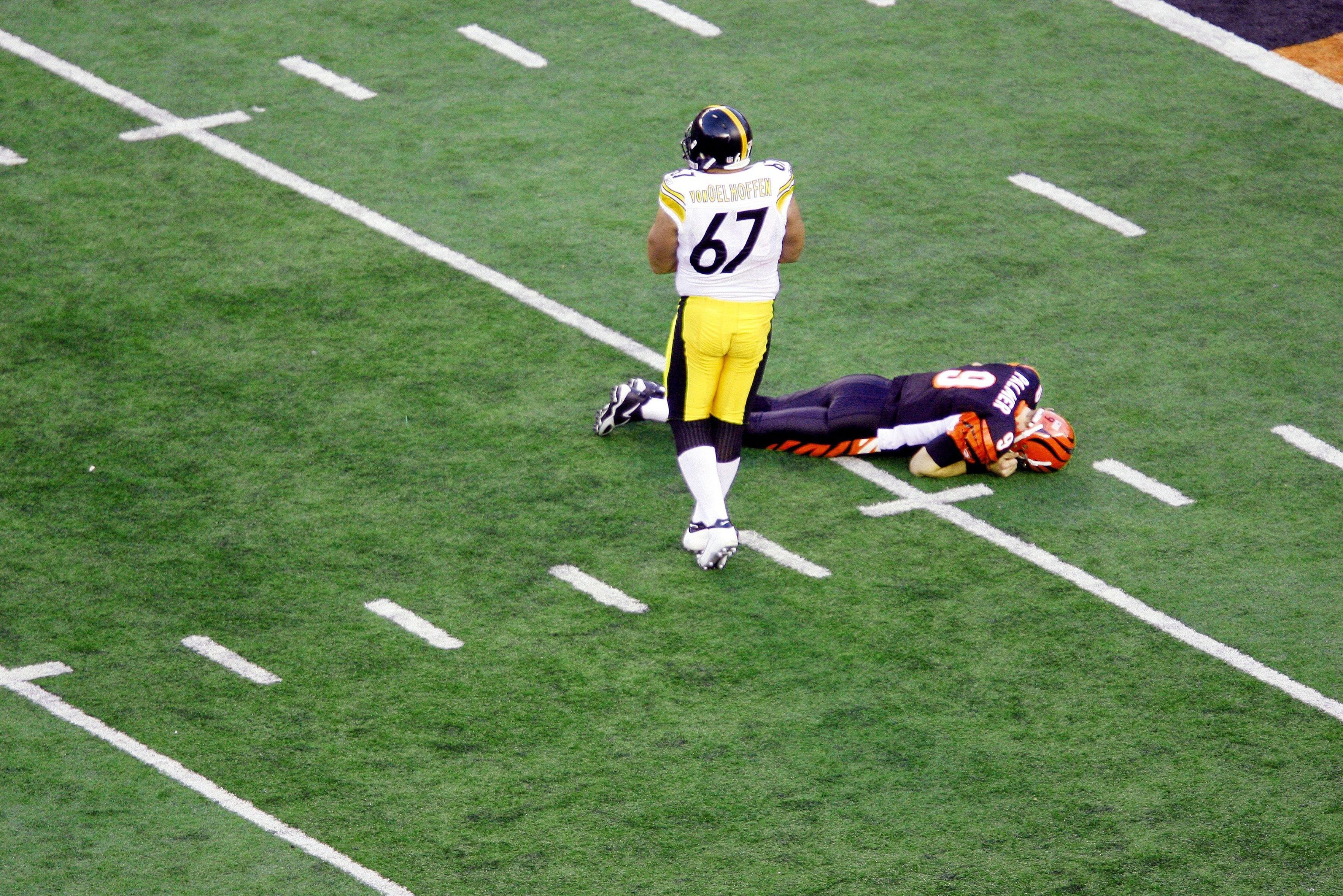 Overhead view of a football field showing a player in a white and yellow uniform with number 67 standing over a downed opponent in orange and black on green turf with white yard lines.