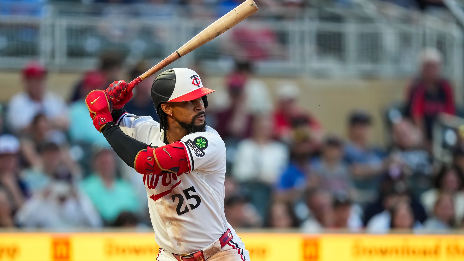 Byron Buxton in white uniform with red trim and red gloves swings a wooden bat. He wears a white helmet with a red and black logo; jersey shows number 25. Stadium crowd blur in the background.