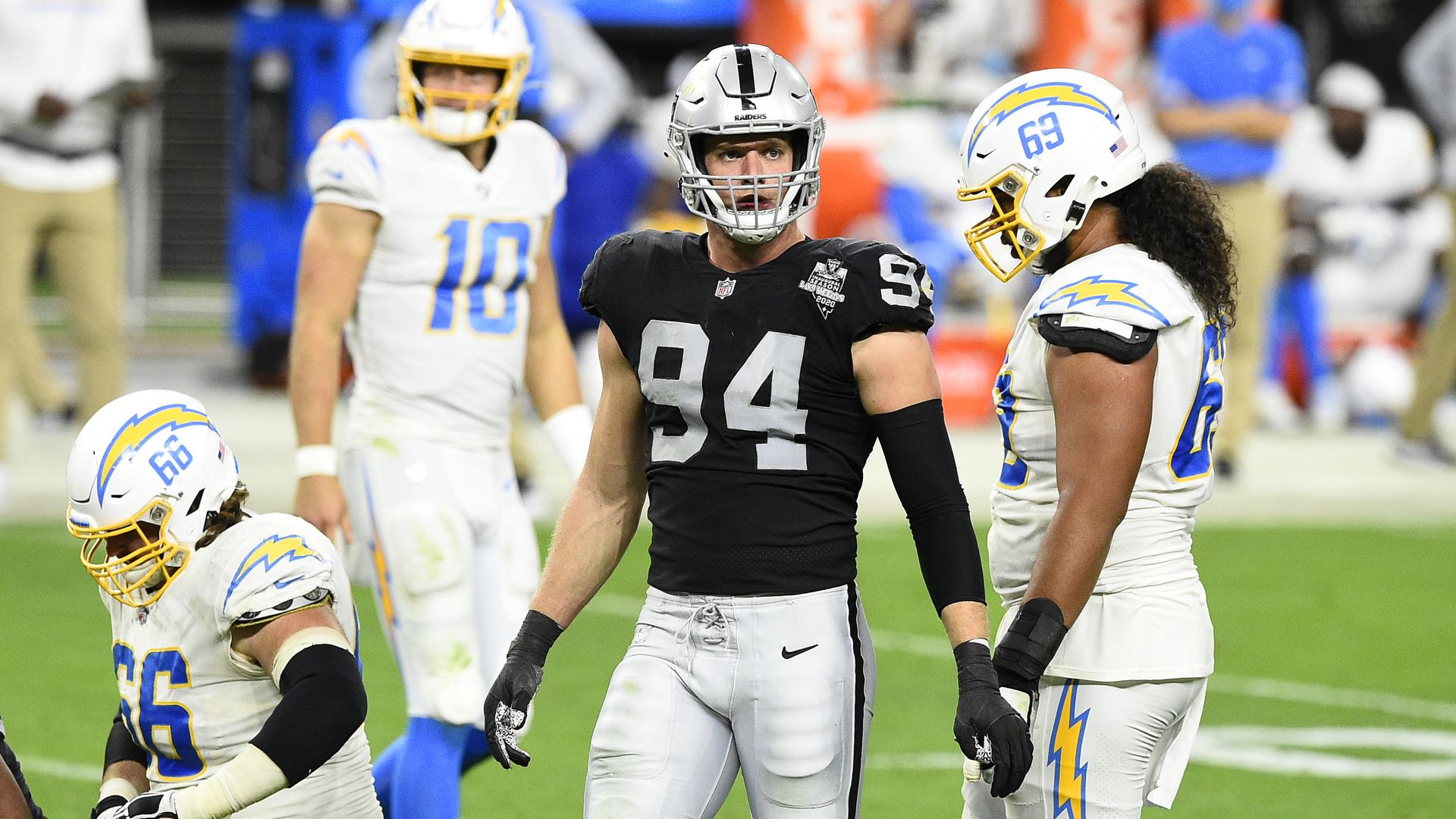 Photo of 4 helmeted football players, one in a black uniform and the other 3 in white uniforms, standing on a field