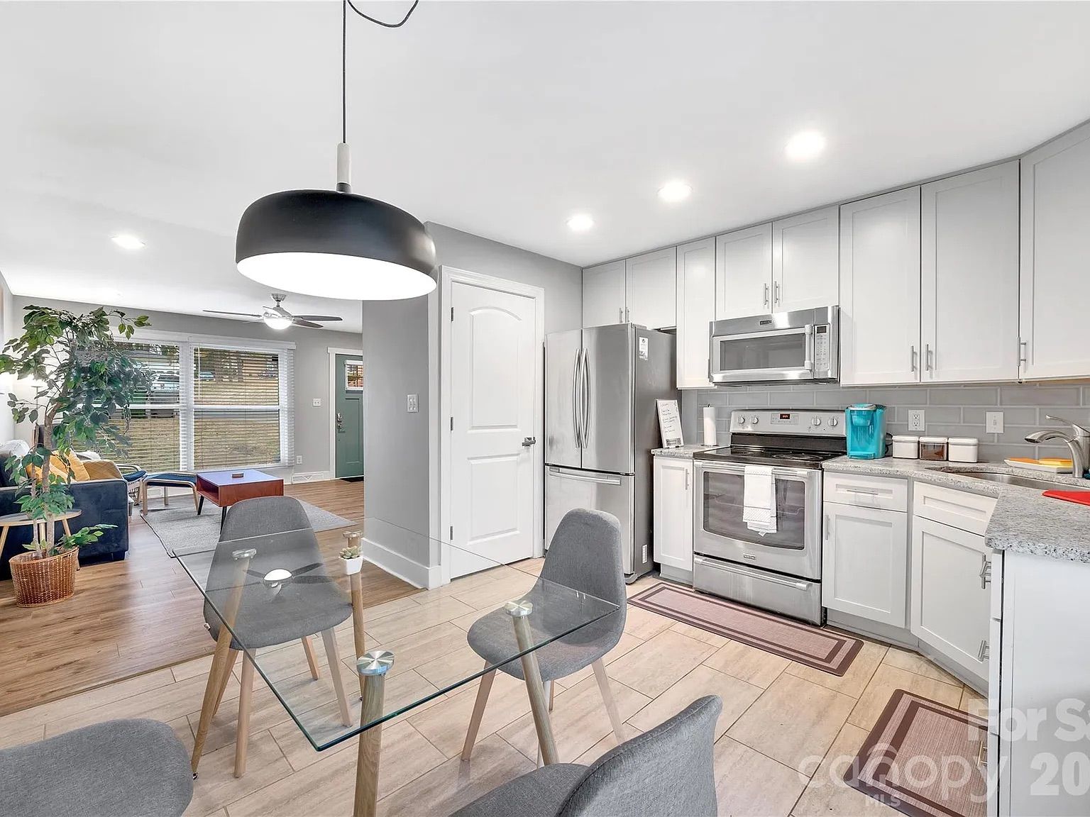 Open-concept kitchen with white cabinets, stainless-steel fridge, oven, and microwave; glass dining table with gray chairs; gray backsplash; living area with a potted plant in view.