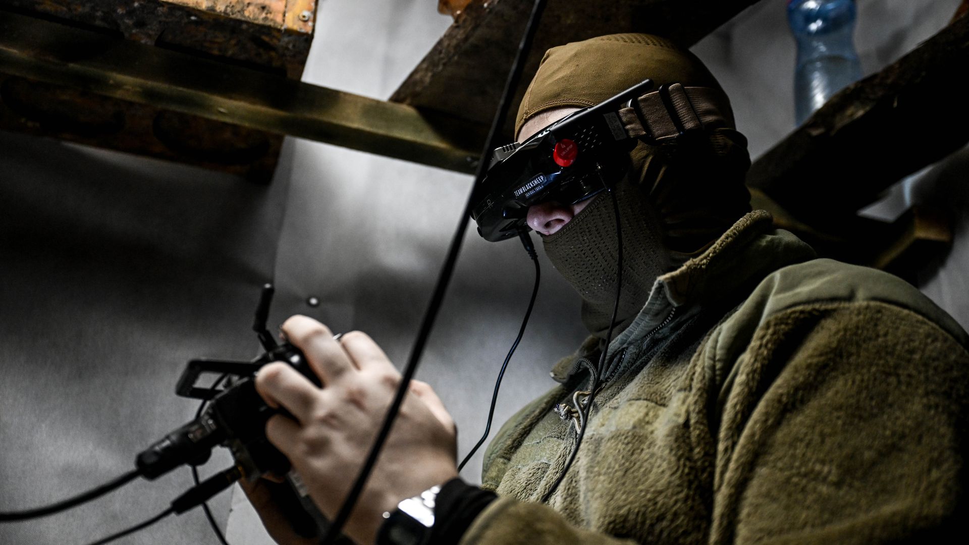 A man is seated, holding a drone controller and wearing a futuristic looking headset. Shelves and items are seen above his head.