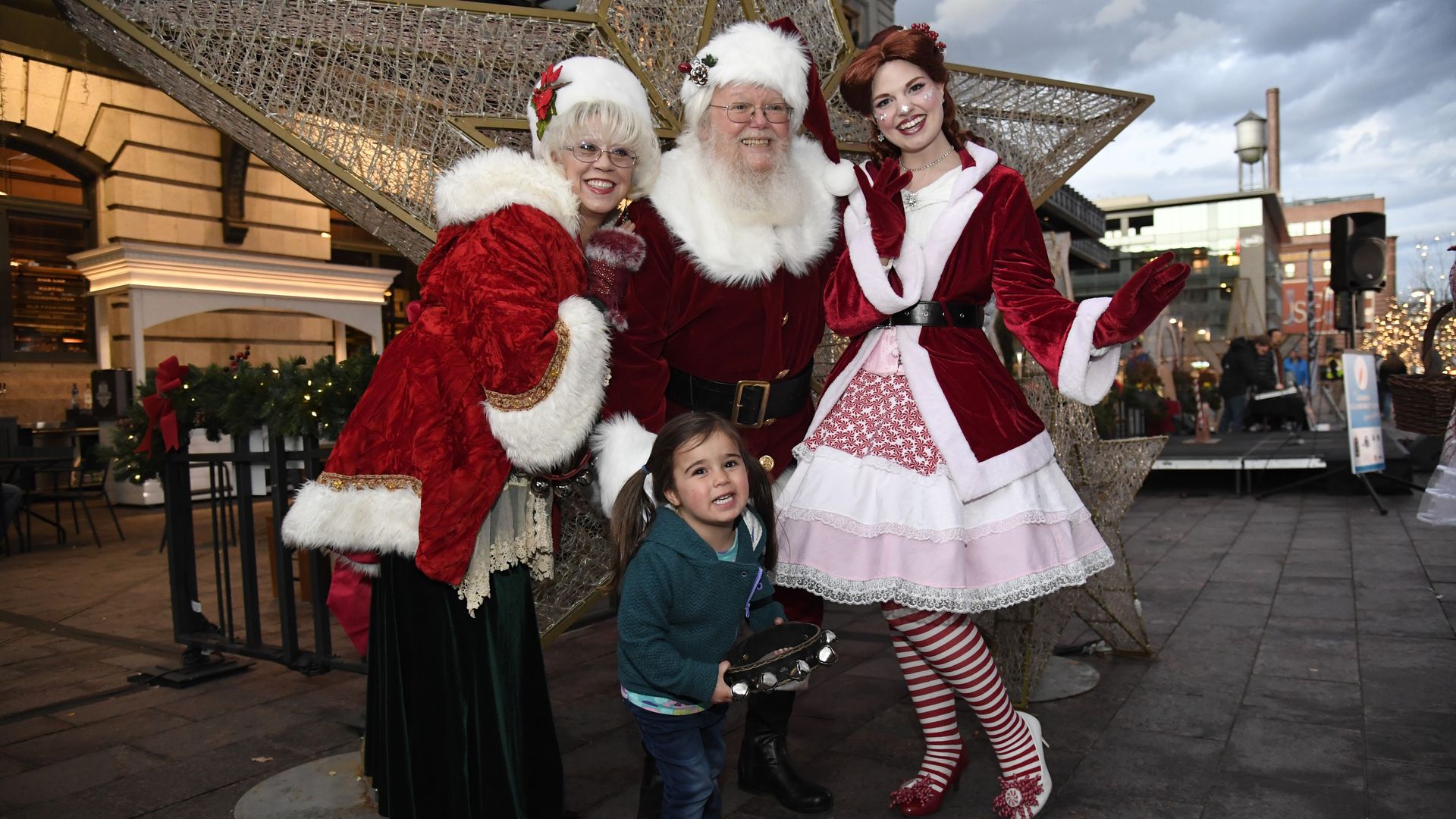 Santa Clause surrounded by Mrs. Clause and a helper next to a child outside. 