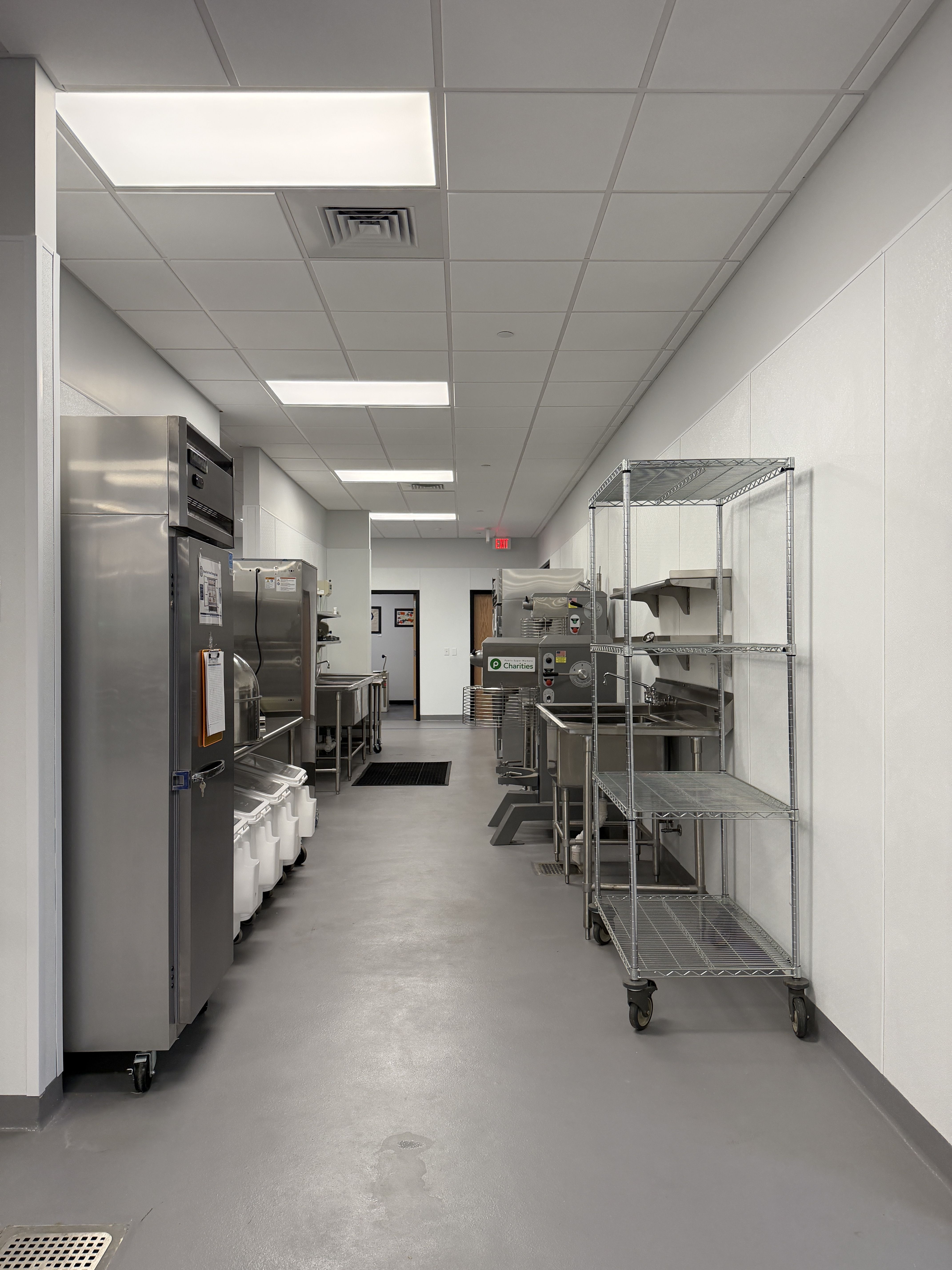 Industrial storage area with silver foil-wrapped containers lined on a wheeled rack beneath white ceiling panels and bright overhead lights; a blue line marks the clean floor.