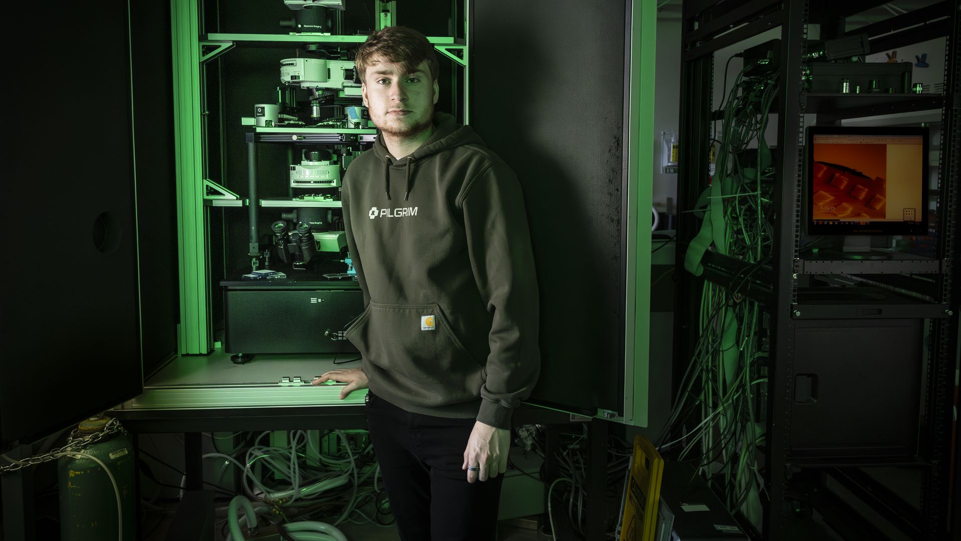 Young man in a dark green "PILGRIM" hoodie stands in a dimly lit lab with green lighting, surrounded by scientific equipment, cables, and a computer showing microscope images.