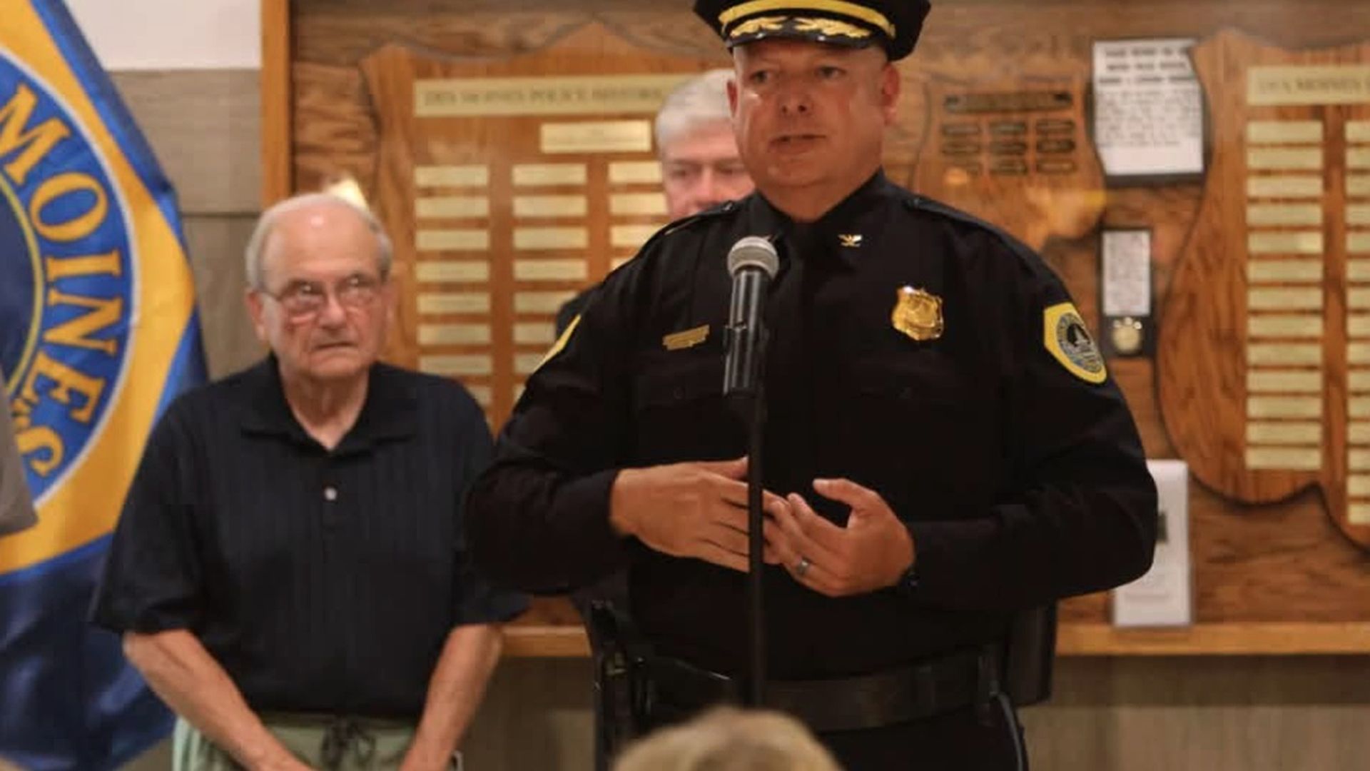 Police officer in a dark uniform speaks into a microphone at a podium, gesturing with his hands. Behind him, an older man watches as a blue-and-gold flag and wood plaques fill the background.
